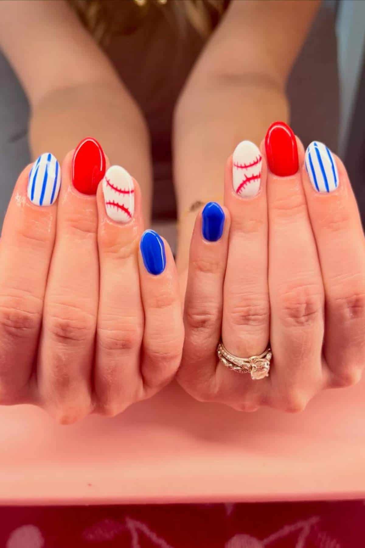 Hands with manicured nails featuring red, blue, and white colors with designs resembling baseballs and pinstripes, shown against a blurred background.