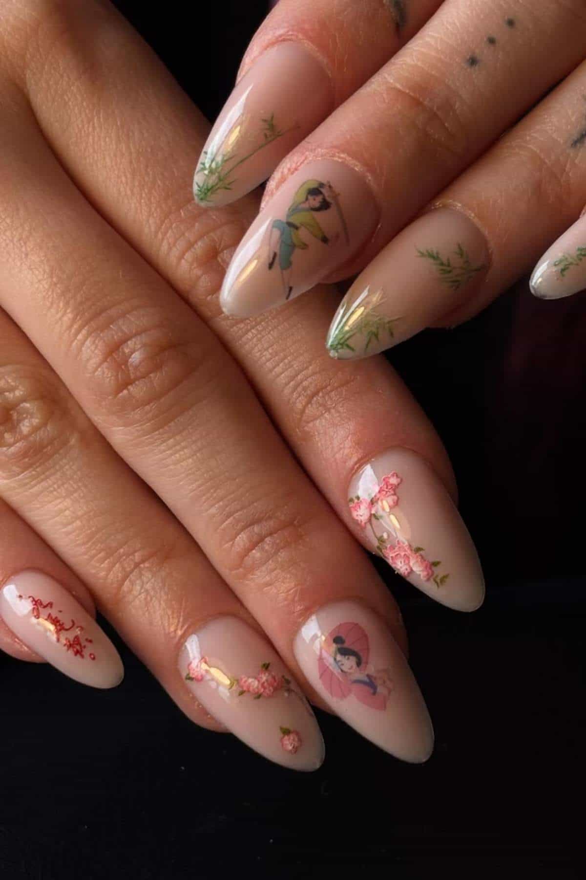 Hands with long, almond-shaped nails featuring nude polish and detailed nail art, including floral designs and small painted figures on several nails.