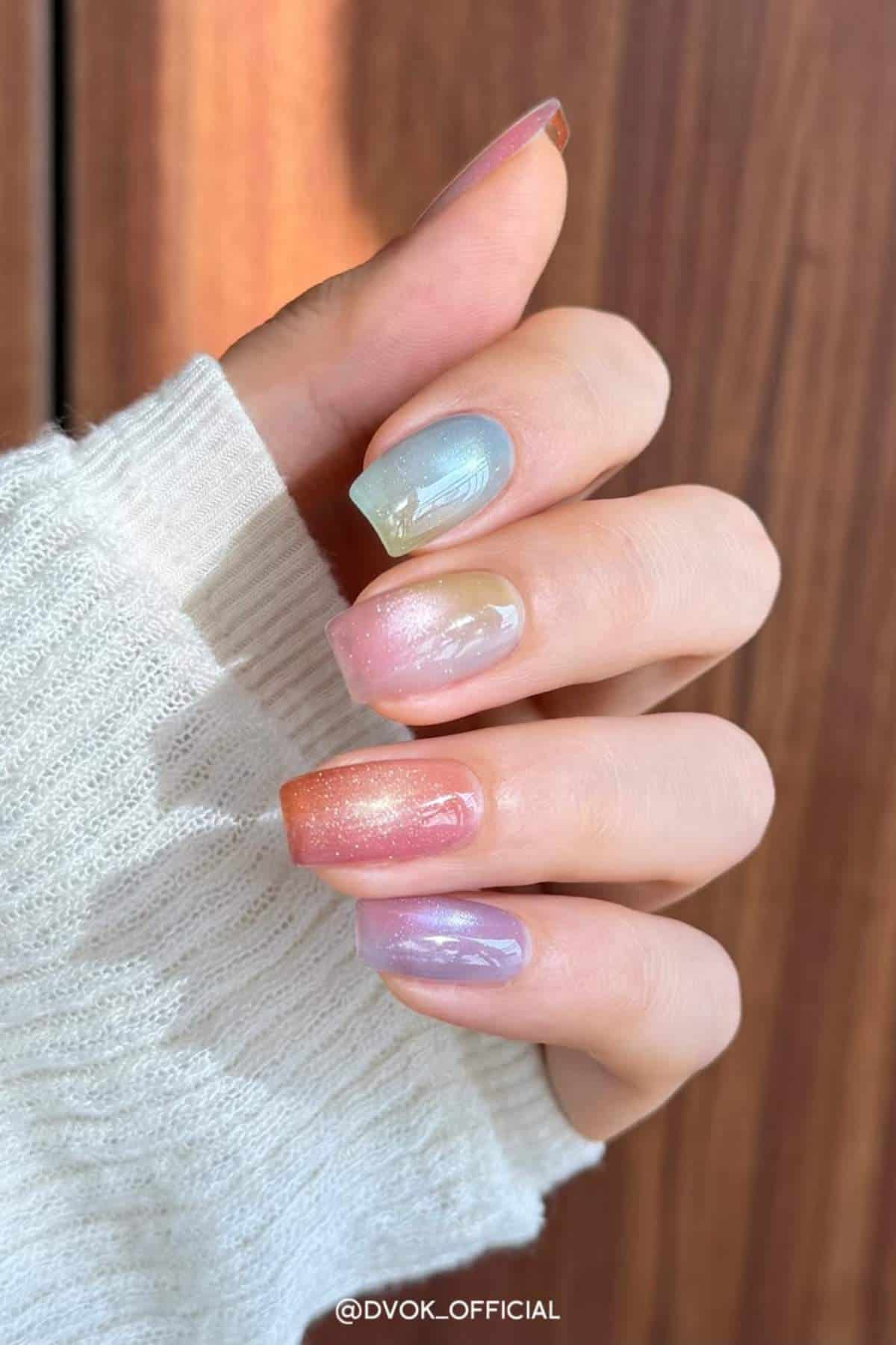 A hand with short, square-shaped nails painted in pastel gradient colors, each nail featuring a different shimmery hue. The person is wearing a light knit sweater.