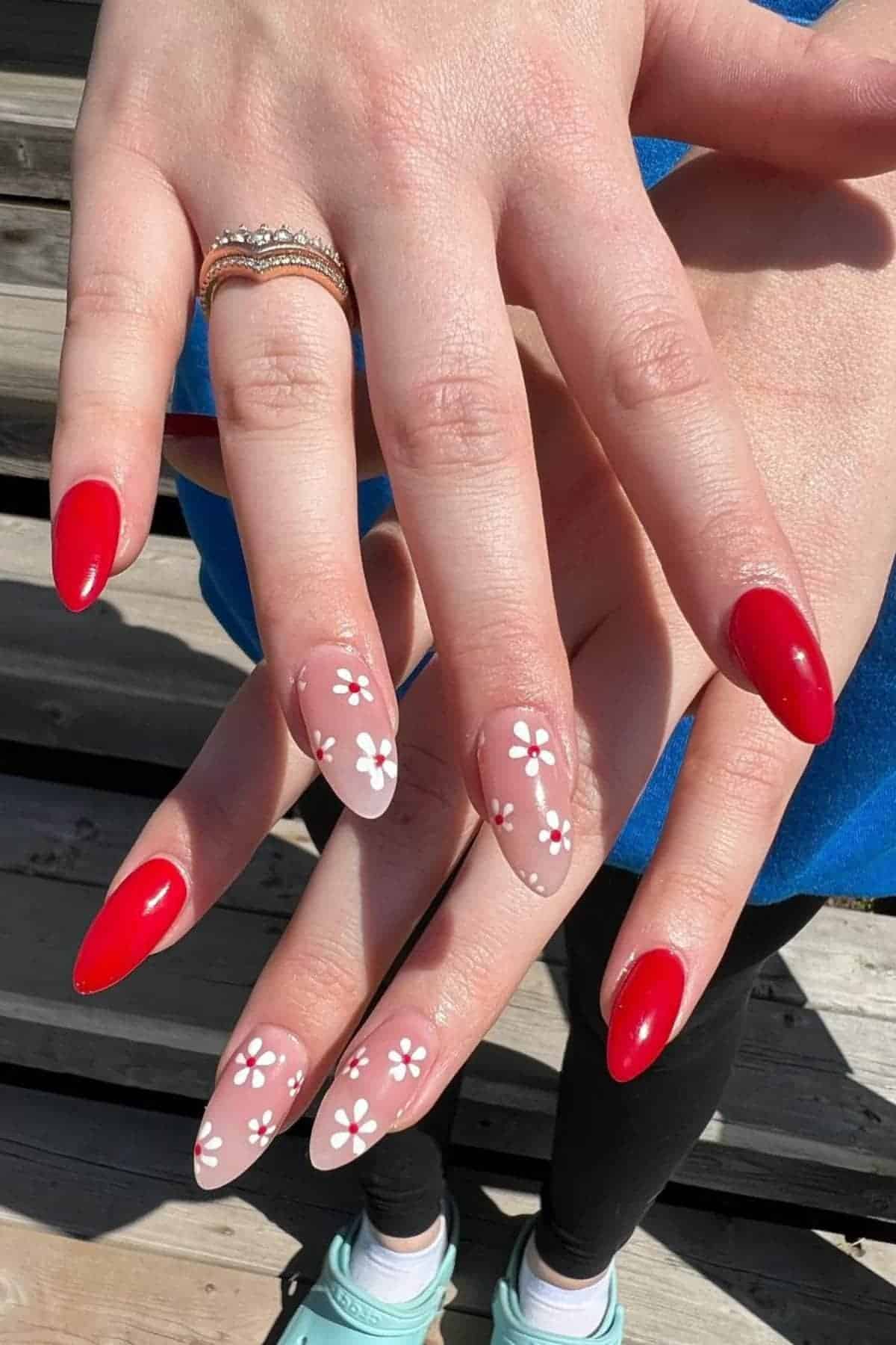 A person with long almond-shaped nails; some nails are painted red, others are nude with white daisy flower designs. The person wears rings and blue clothing.