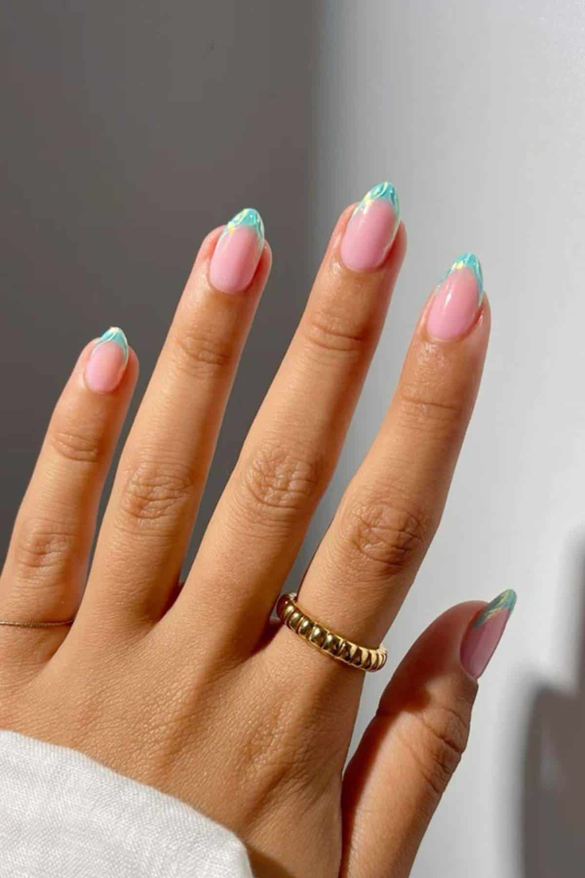A hand with a gold ring displays nails painted with a pink base and light blue-green wavy French tips against a neutral background.