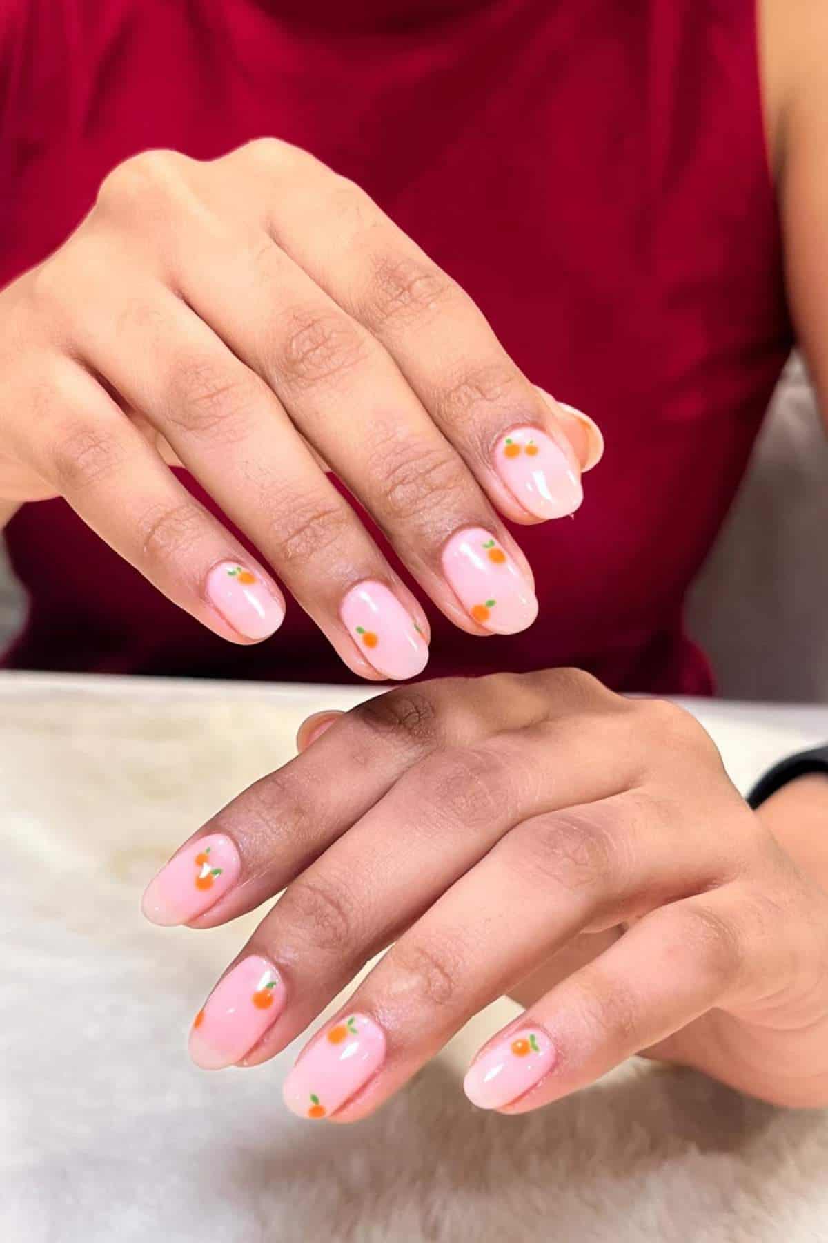 Close-up of hands with short, almond-shaped nails featuring a glossy light pink base and small orange and green floral nail art.