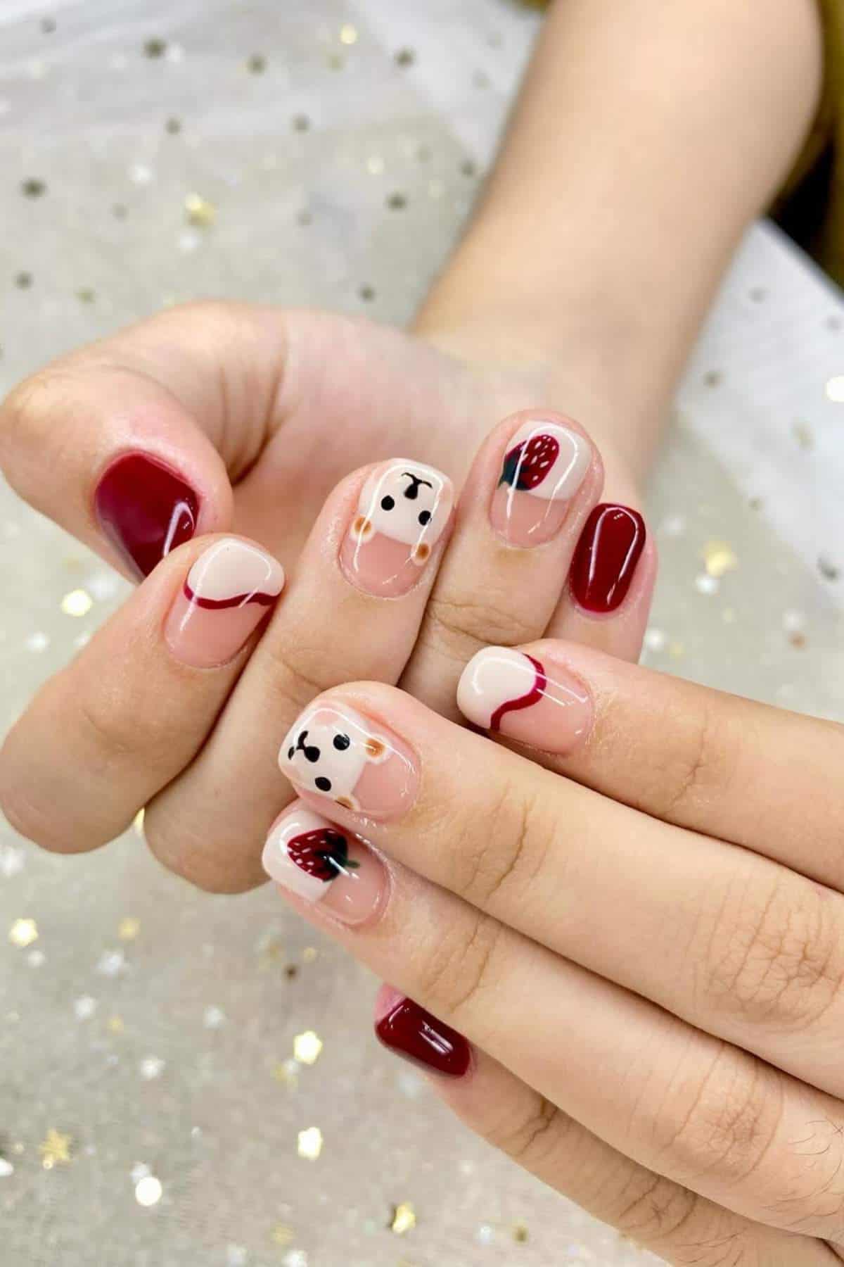 A hand with short nails painted in a mix of burgundy, nude, and white polish, featuring abstract designs and black dot patterns on a sheer, star-patterned fabric background.