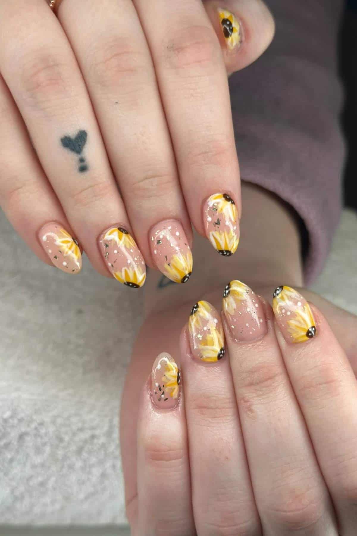 Hands with manicured nails featuring yellow sunflower designs, black tips, and small glitter accents; one finger has a small heart tattoo.