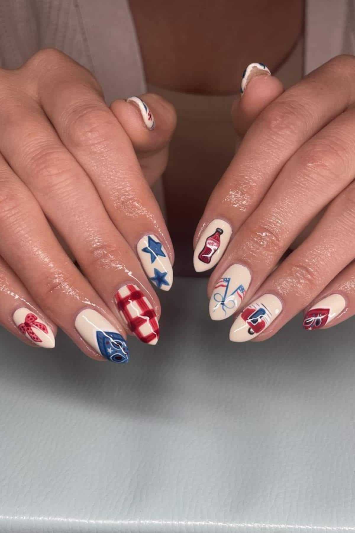 Close-up of hands with long nails painted in a white base and decorated with red, blue, and patriotic-themed designs, including stars, stripes, sunglasses, and bottles.