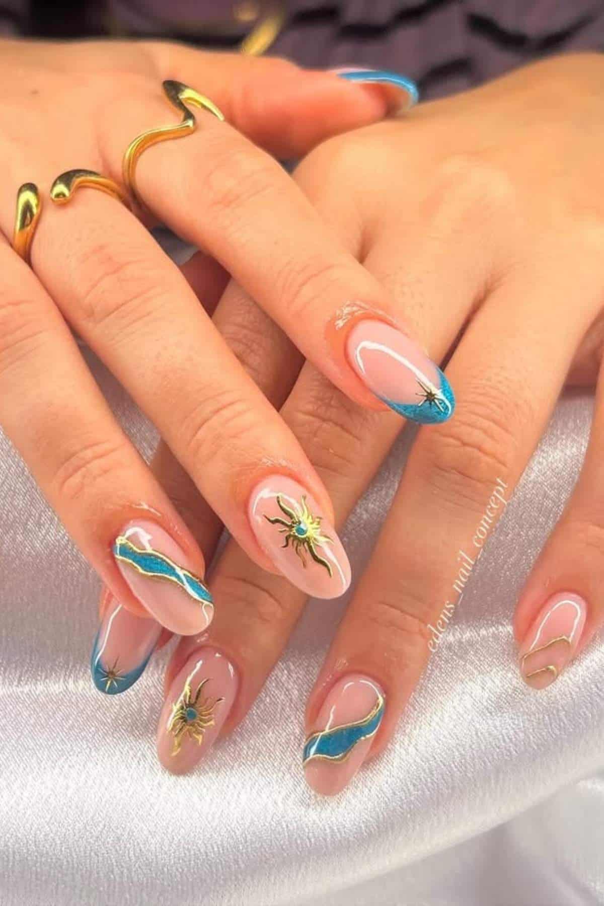 Hands with long, almond-shaped nails featuring blue and gold abstract tips and small, detailed sun designs on a few nails. The person is wearing gold rings.