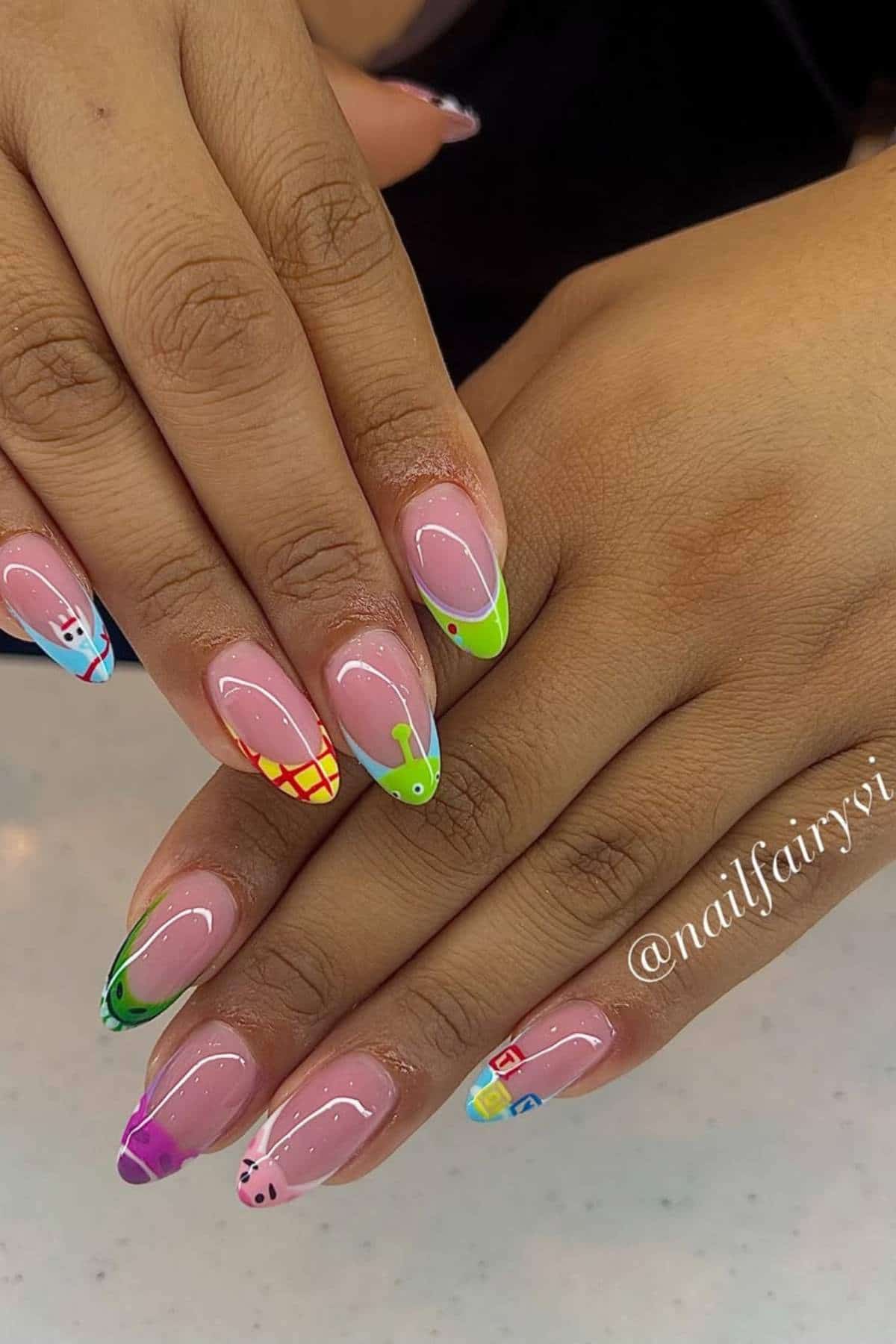 Hands with almond-shaped nails featuring colorful and playful cartoon designs, including a fish, ladybug, grid pattern, and other decorative elements.