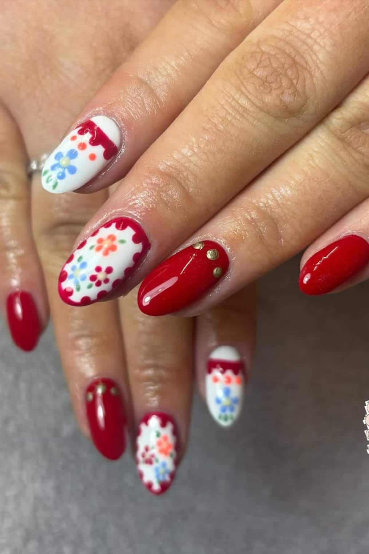 Close-up of hands with almond-shaped nails featuring red polish, floral nail art on white accent nails, and gold stud accents.