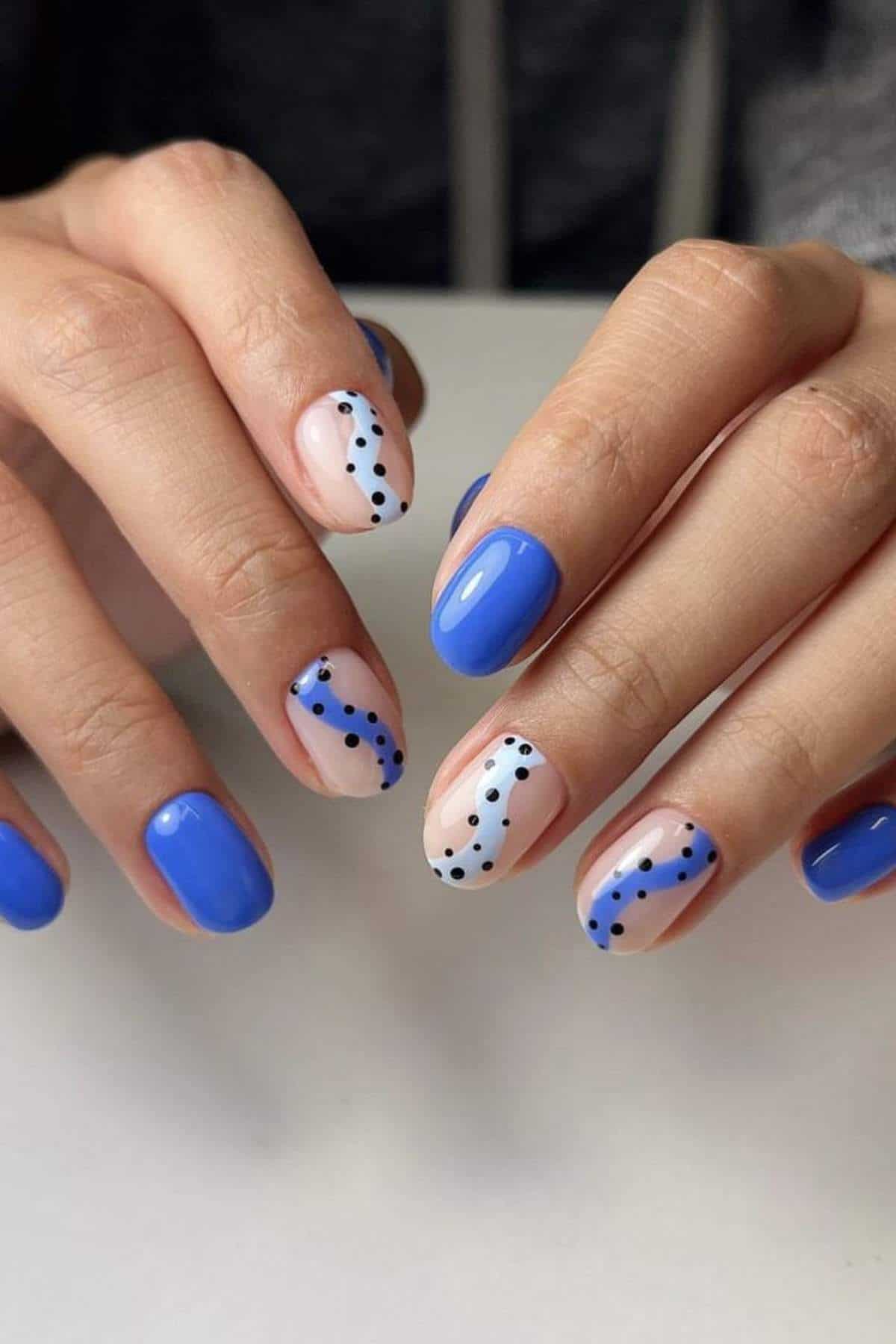 Hands with short nails painted blue; two nails on each hand have a nude base with blue waves and black dots as accents.