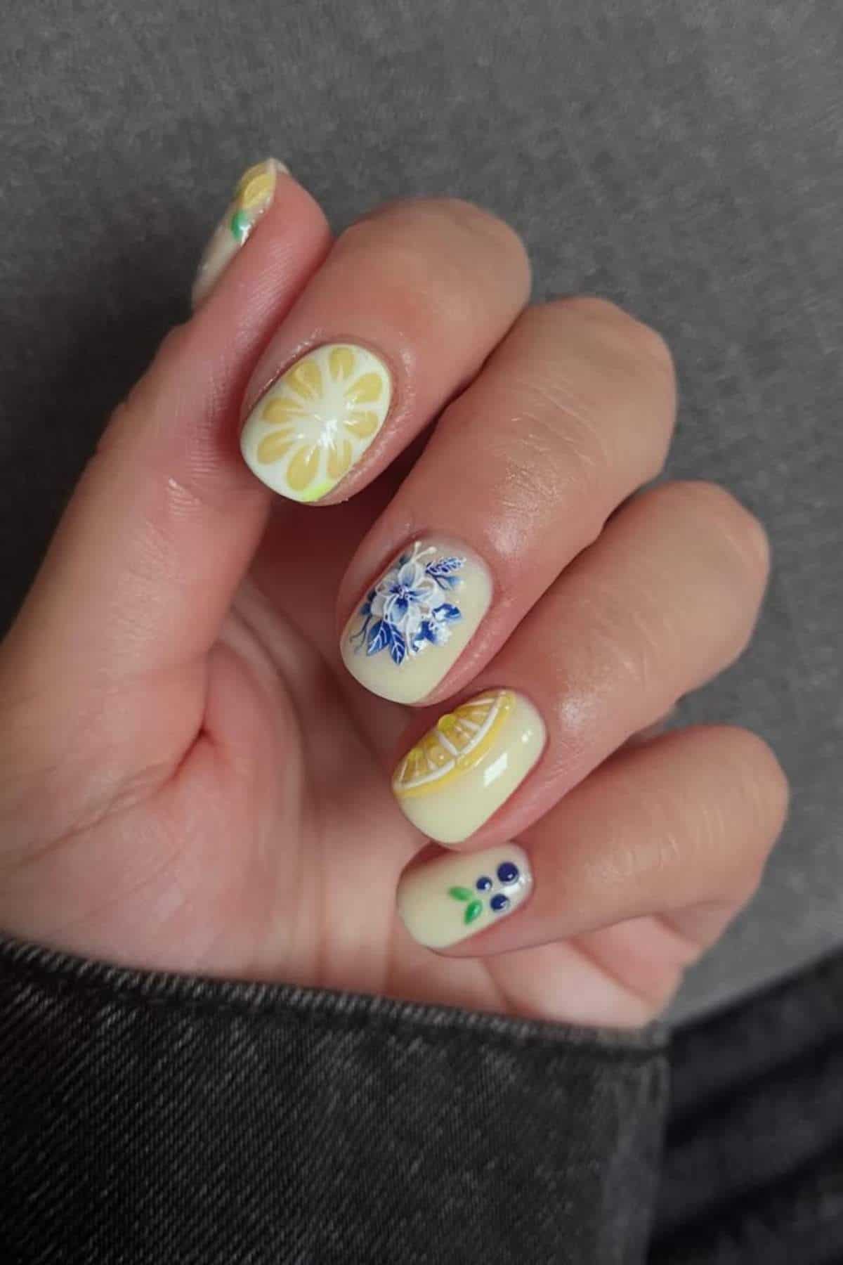 A hand with yellow-themed nail art featuring lemon slices, a blue floral design, and blue berries on a light background.