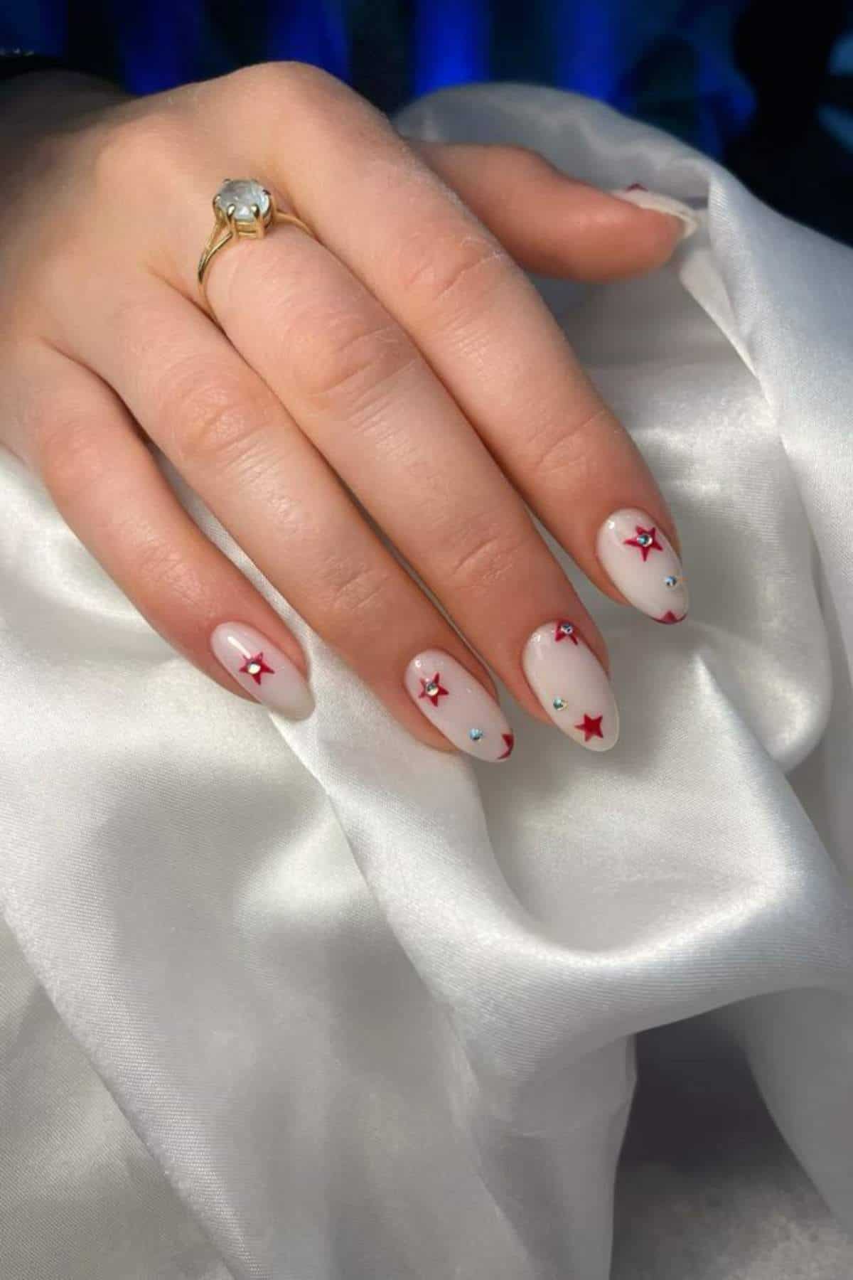 A hand with almond-shaped nails painted pale pink, decorated with red star stickers and small rhinestones, rests on white satin fabric. The person wears a gold ring with a clear gemstone.