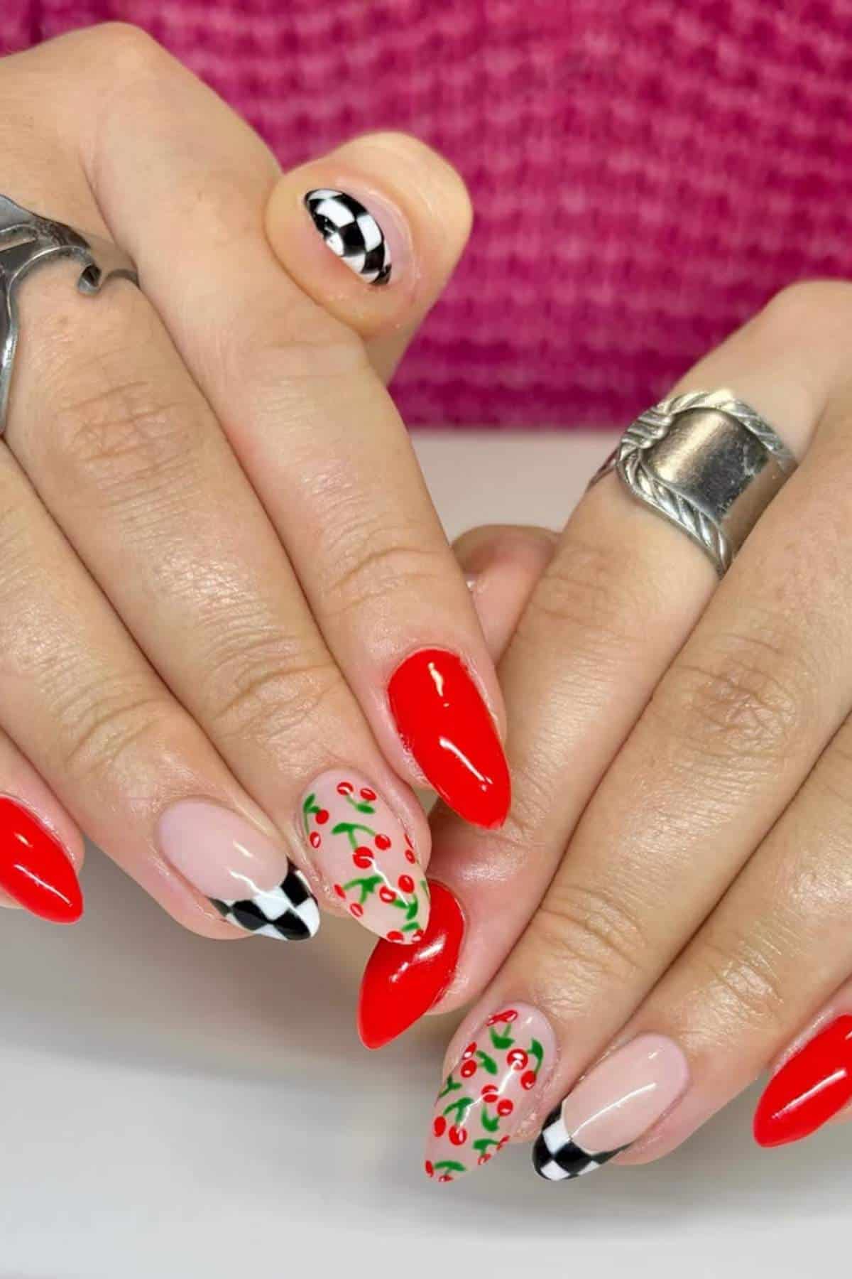 Hands with long almond-shaped nails featuring a mix of designs: red polish, black-and-white checkered tips, and nude nails with small red and green cherry patterns.