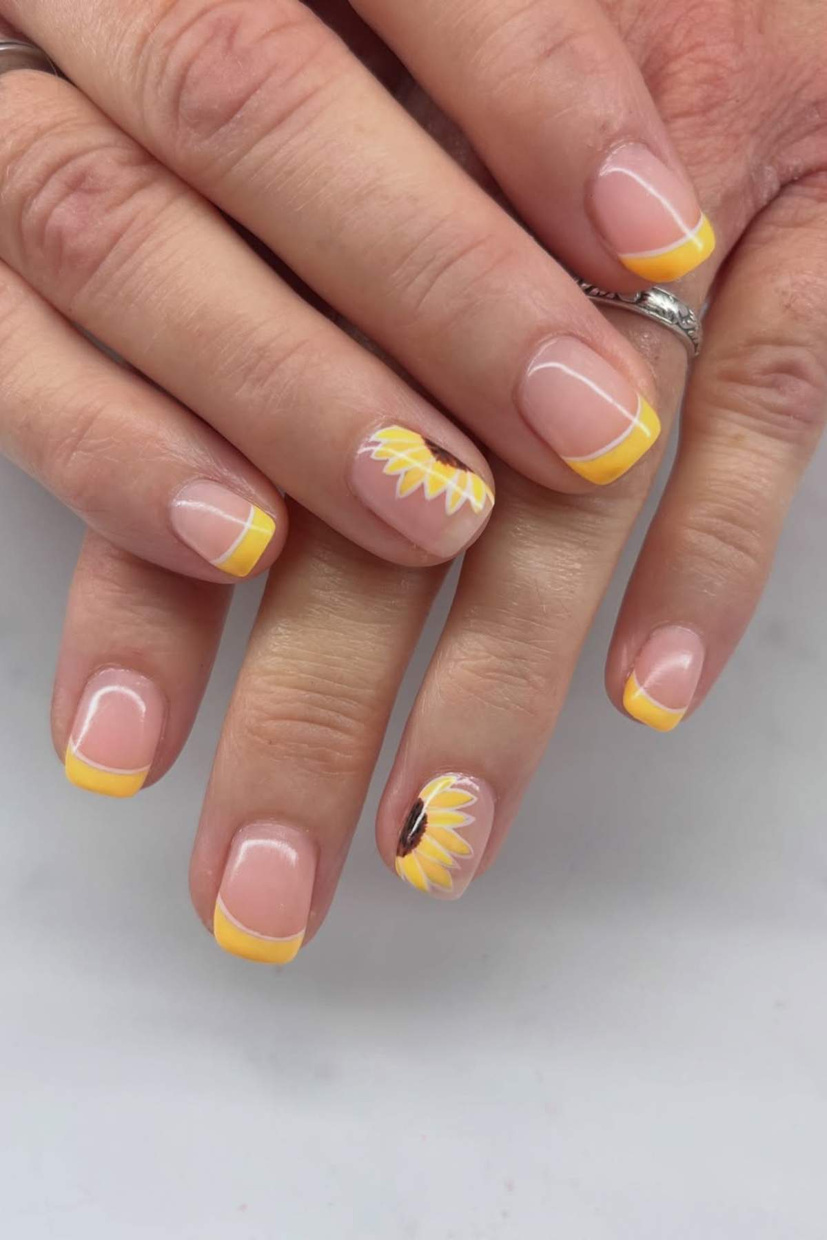 Hands with short, square-shaped nails featuring yellow French tips; the ring fingernail has a sunflower design with yellow petals and a brown center.