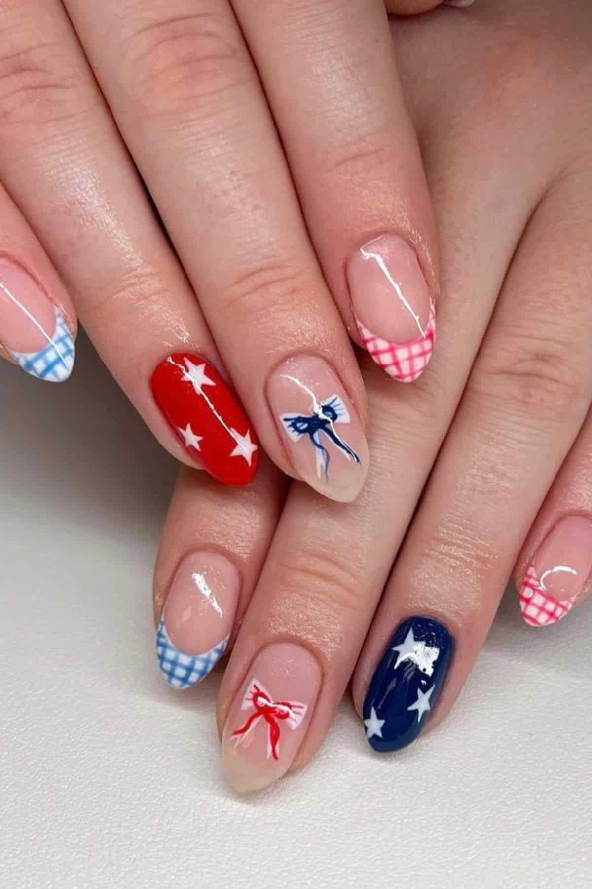 Hands with manicured nails featuring red, white, and blue designs, including stars, bows, and gingham patterns on an almond-shaped nail style.
