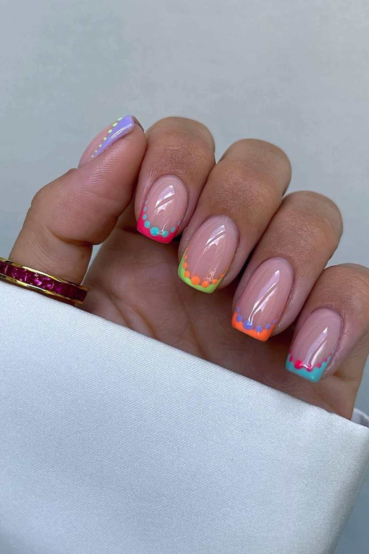 A hand with manicured nails featuring clear polish and colorful, dotted tips in orange, green, blue, and purple, partially covered by a white sleeve.