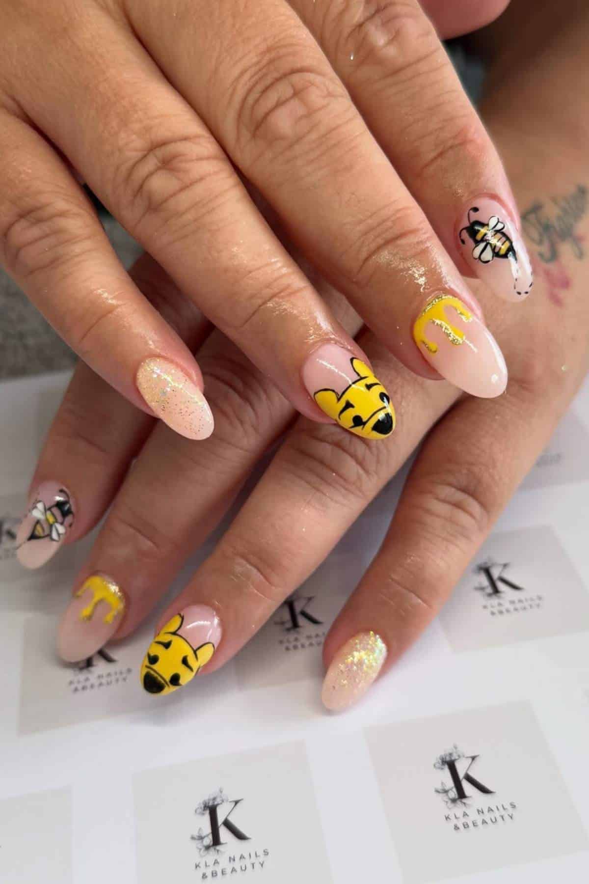 Hands with long almond-shaped nails featuring nude polish, glitter, and bee-themed nail art including yellow and black bees and honey drips.