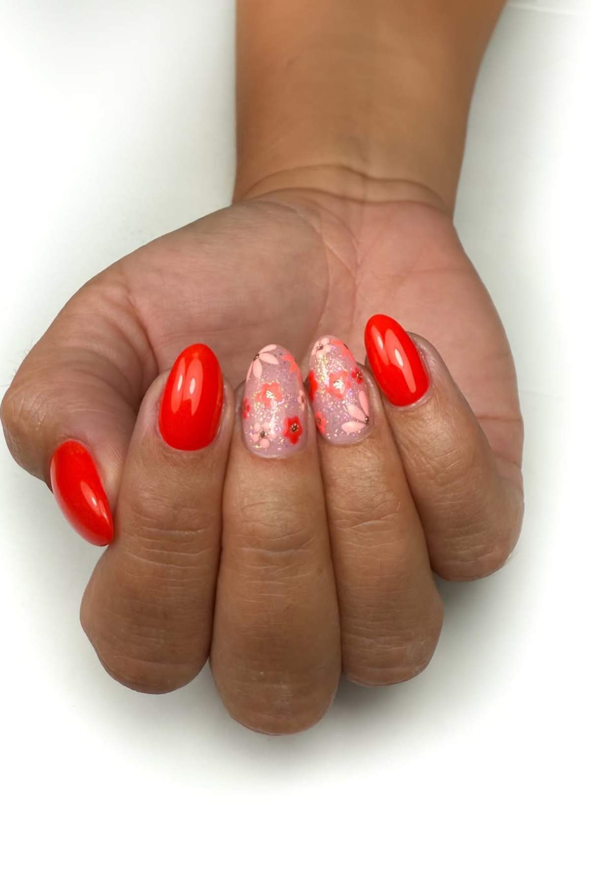 A hand with red polished nails and two accent nails featuring pink floral nail art with glitter and red dots.
