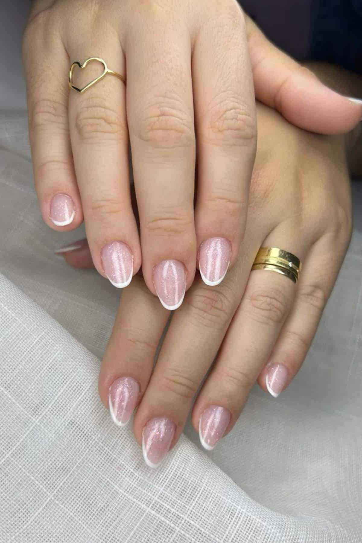 Hands with gold rings rest on a white surface, displaying short nails with a light pink base and white French tips.