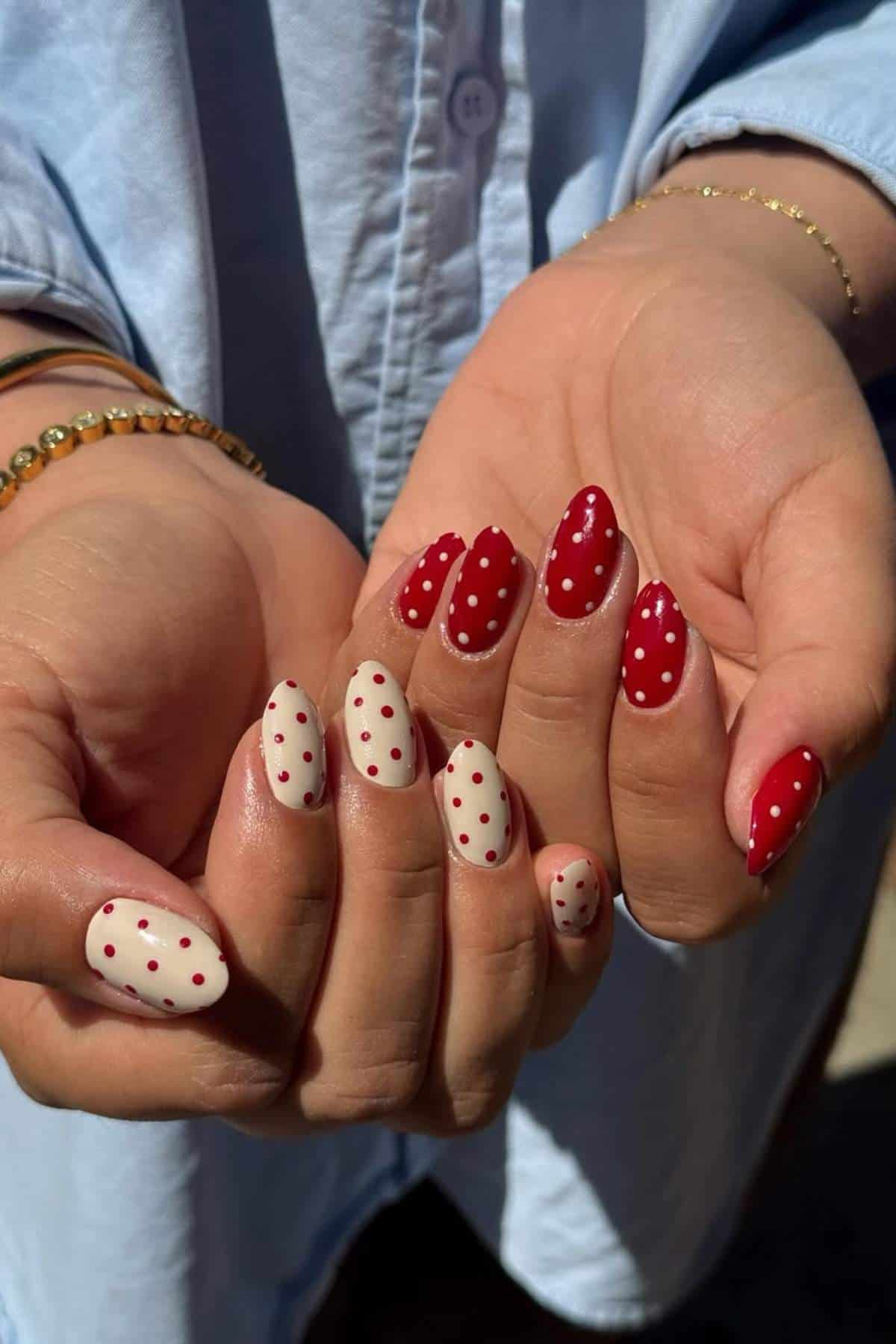 Hands with manicured nails; some nails are painted red with white polka dots, others are white with red polka dots. Person is wearing a light blue shirt and gold bracelets.