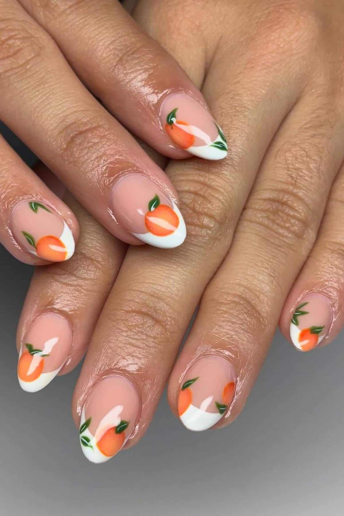Close-up of hands with almond-shaped nails featuring a peach fruit design, white French tips, and green leaf accents on a nude base.