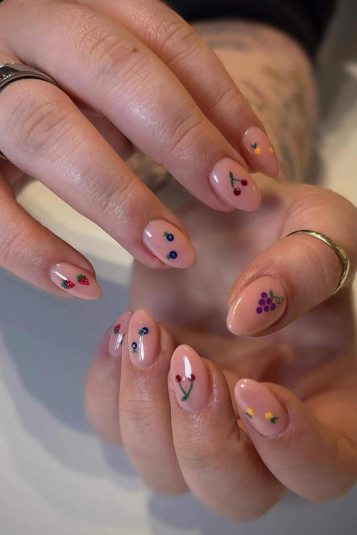 Hands with almond-shaped nails painted nude, each nail decorated with small, colorful fruit designs such as cherries, strawberries, and grapes.
