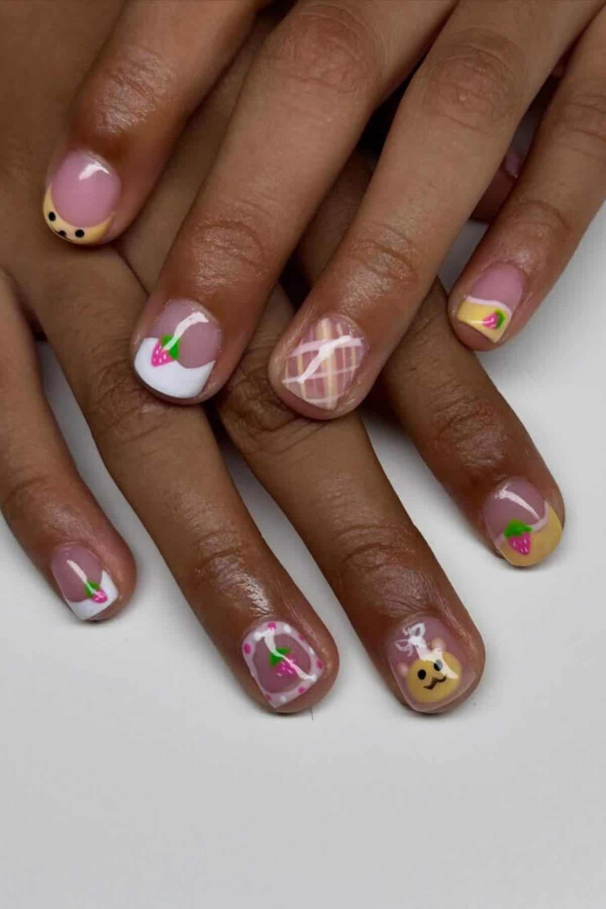 Hands with short nails painted with a variety of colorful designs, including strawberries, plaid, bear faces, and geometric shapes on a white background.