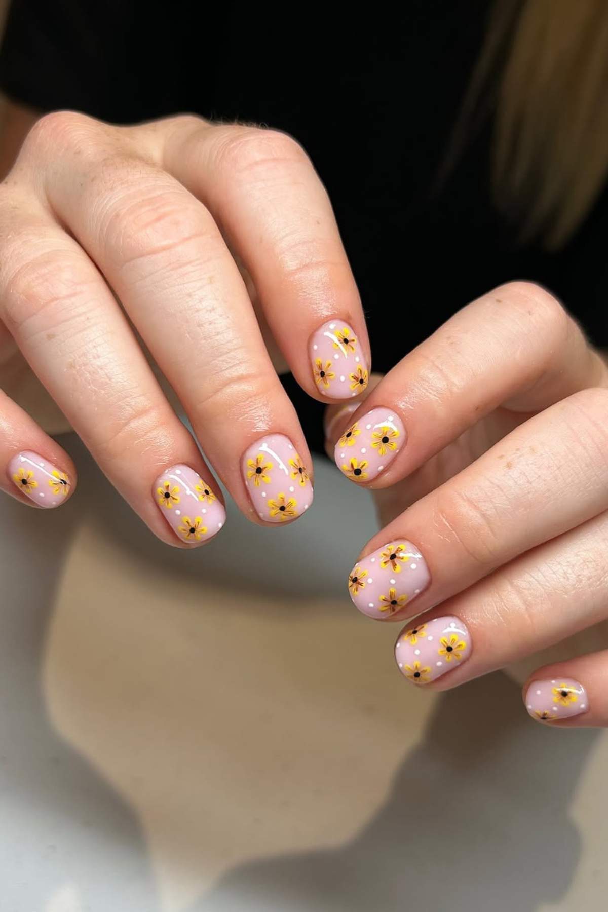 Hands with short, oval-shaped nails painted light pink and decorated with small yellow and white flower designs on each nail.