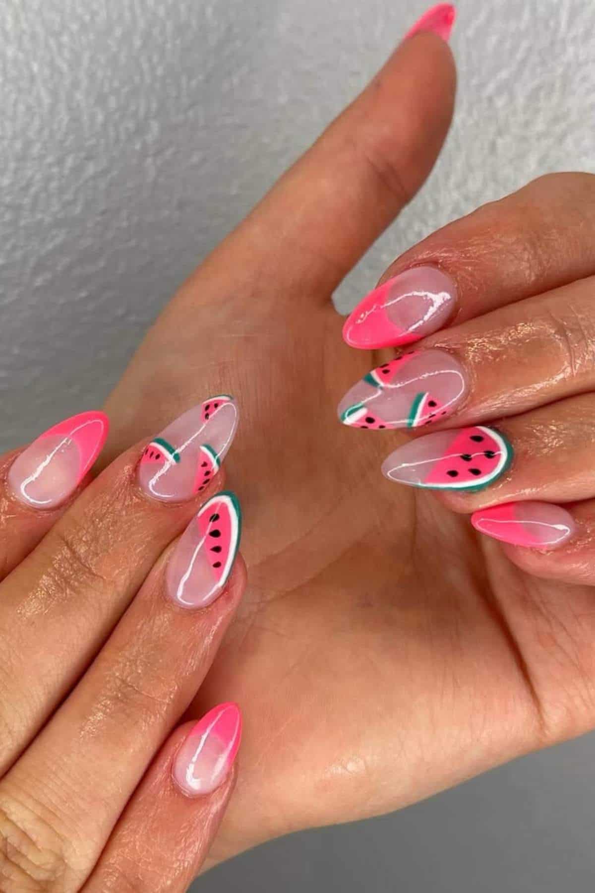 A hand displays almond-shaped nails with a watermelon design, featuring pink tips, green accents, and black seed details over a translucent base.