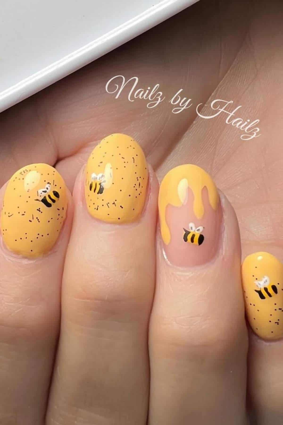 Close-up of hand with yellow nail art featuring small bee designs, black speckles, and a honey drip pattern on one nail.