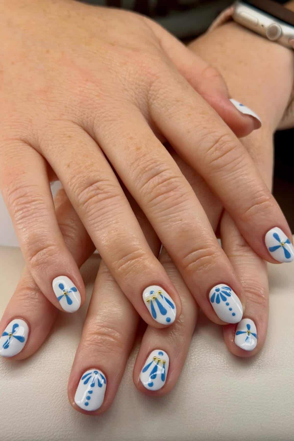 Hands with white nail polish featuring blue, yellow, and dark blue abstract floral and dot designs on short, rounded nails.