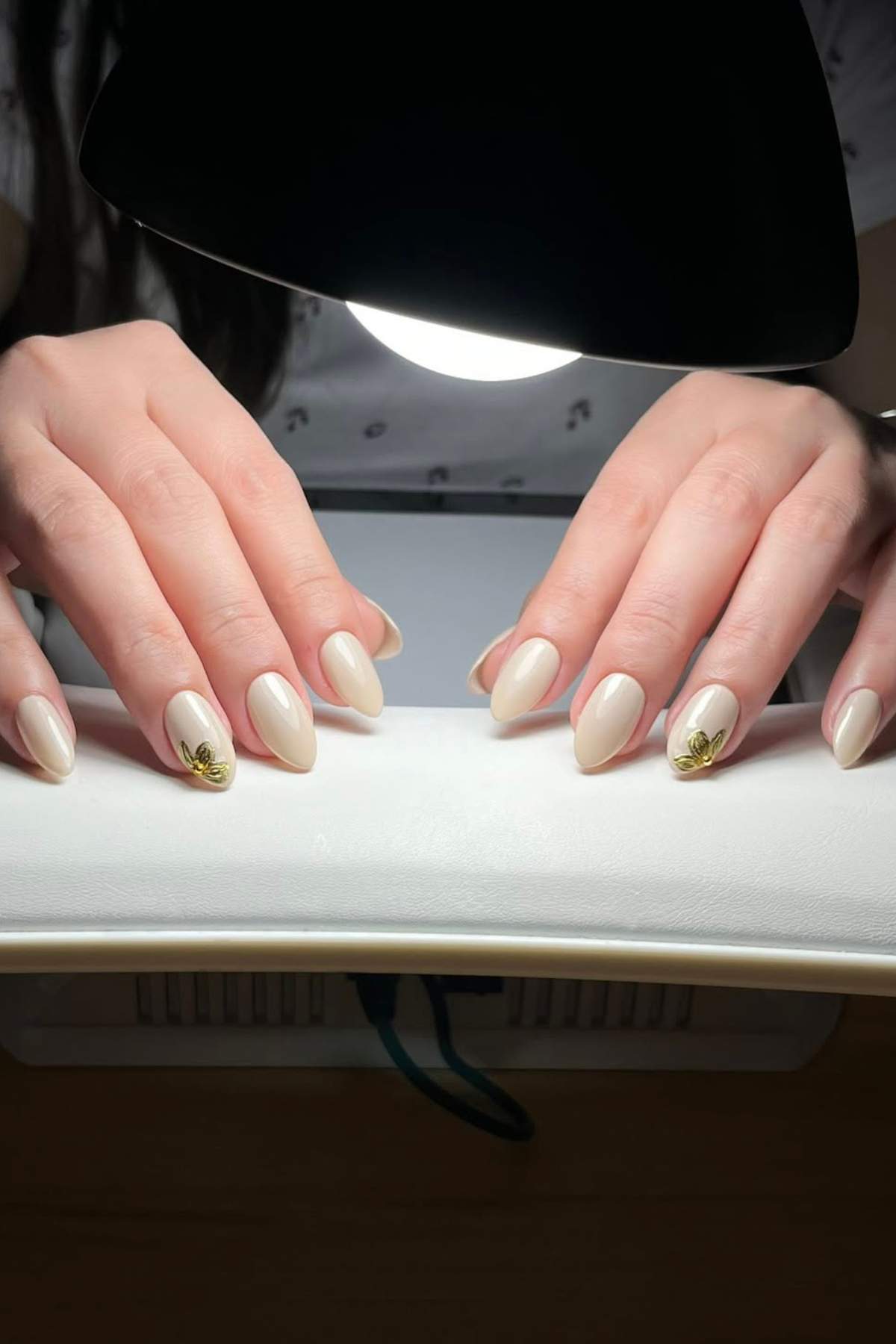 Two hands with almond-shaped, nude-colored nails, each featuring a small gold leaf design on one ring finger, resting under a nail dryer.