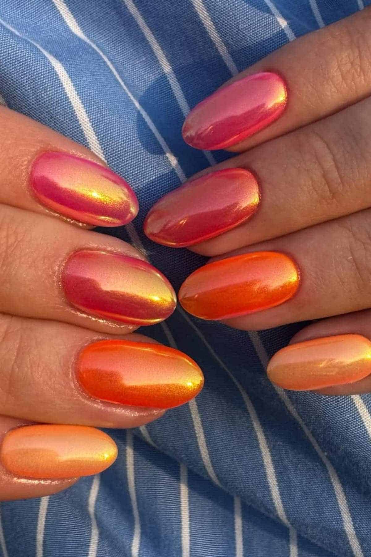 Close-up of hands with oval-shaped nails featuring a gradient manicure in shades of pink, orange, and peach, over a blue and white striped fabric background.