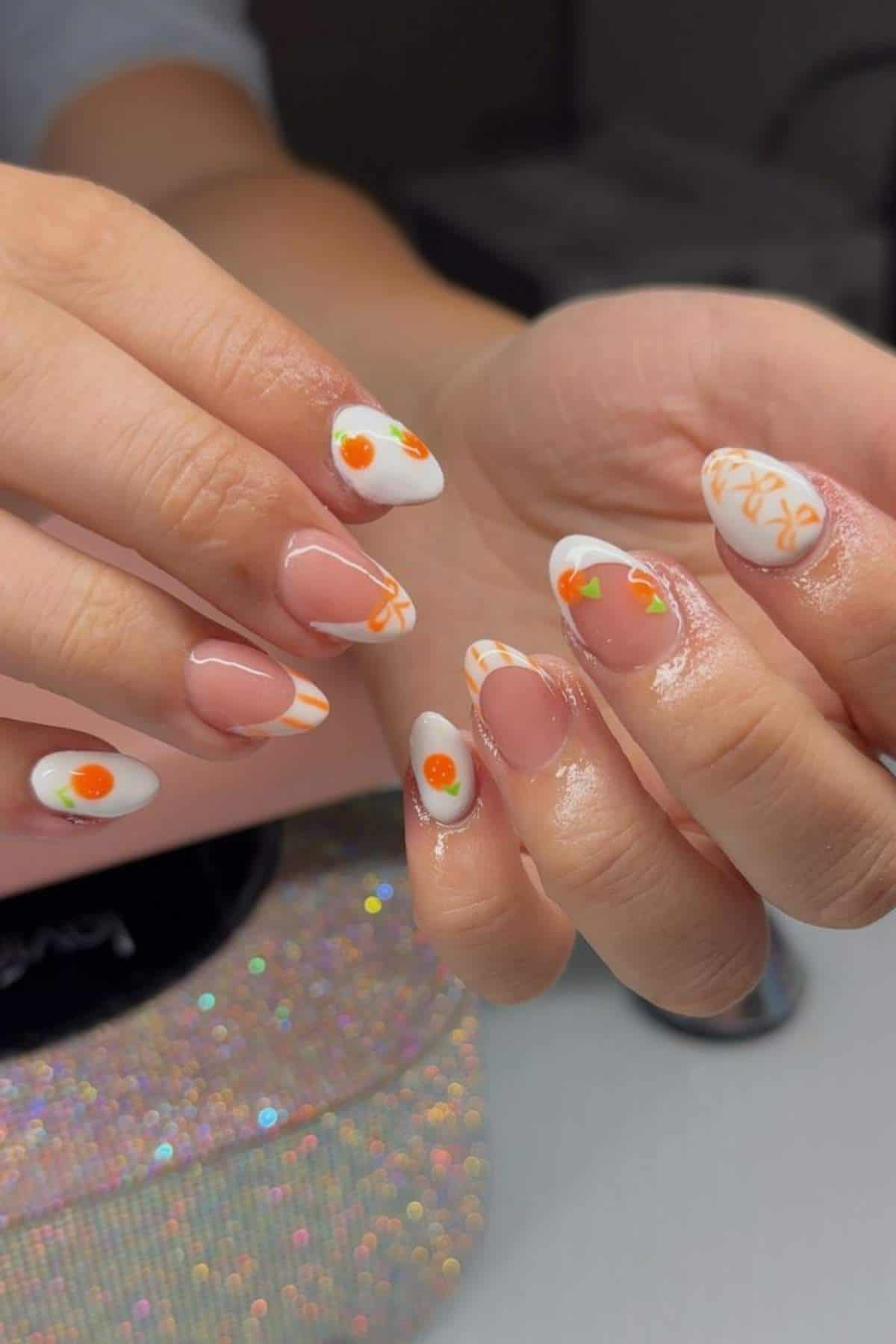 Hands with almond-shaped nails featuring white and clear polish, decorated with orange fruit and leaf designs, and orange squiggly patterns.