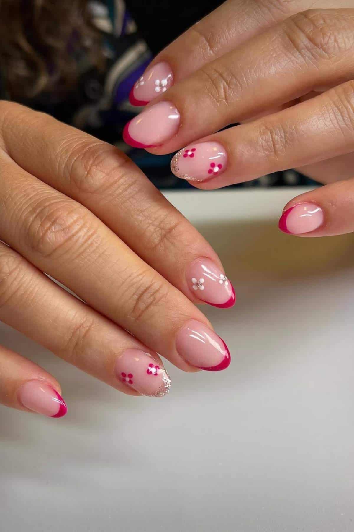 Hands with oval-shaped nails featuring pink French tips and small floral designs on a light nude base.