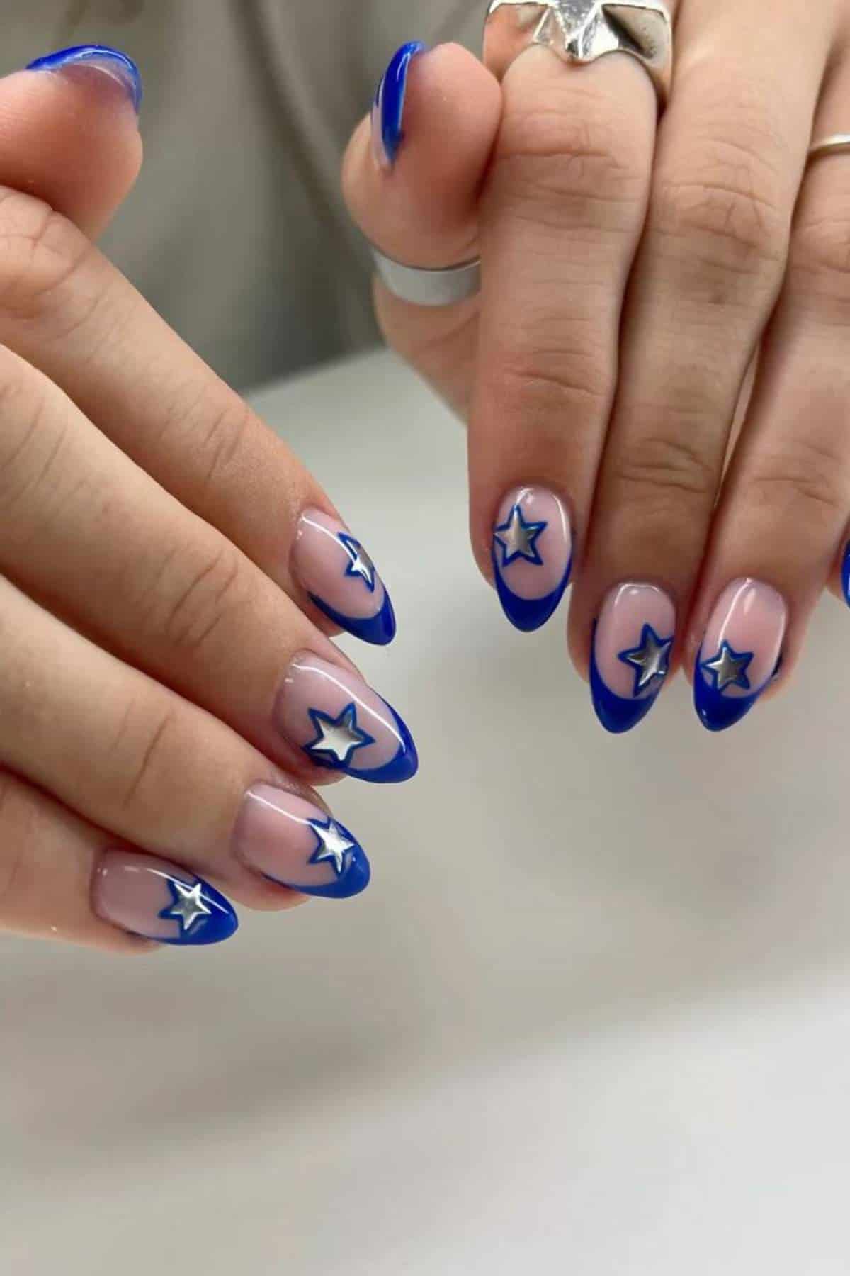 Hands showing almond-shaped nails with blue French tips and silver star designs on a nude base.