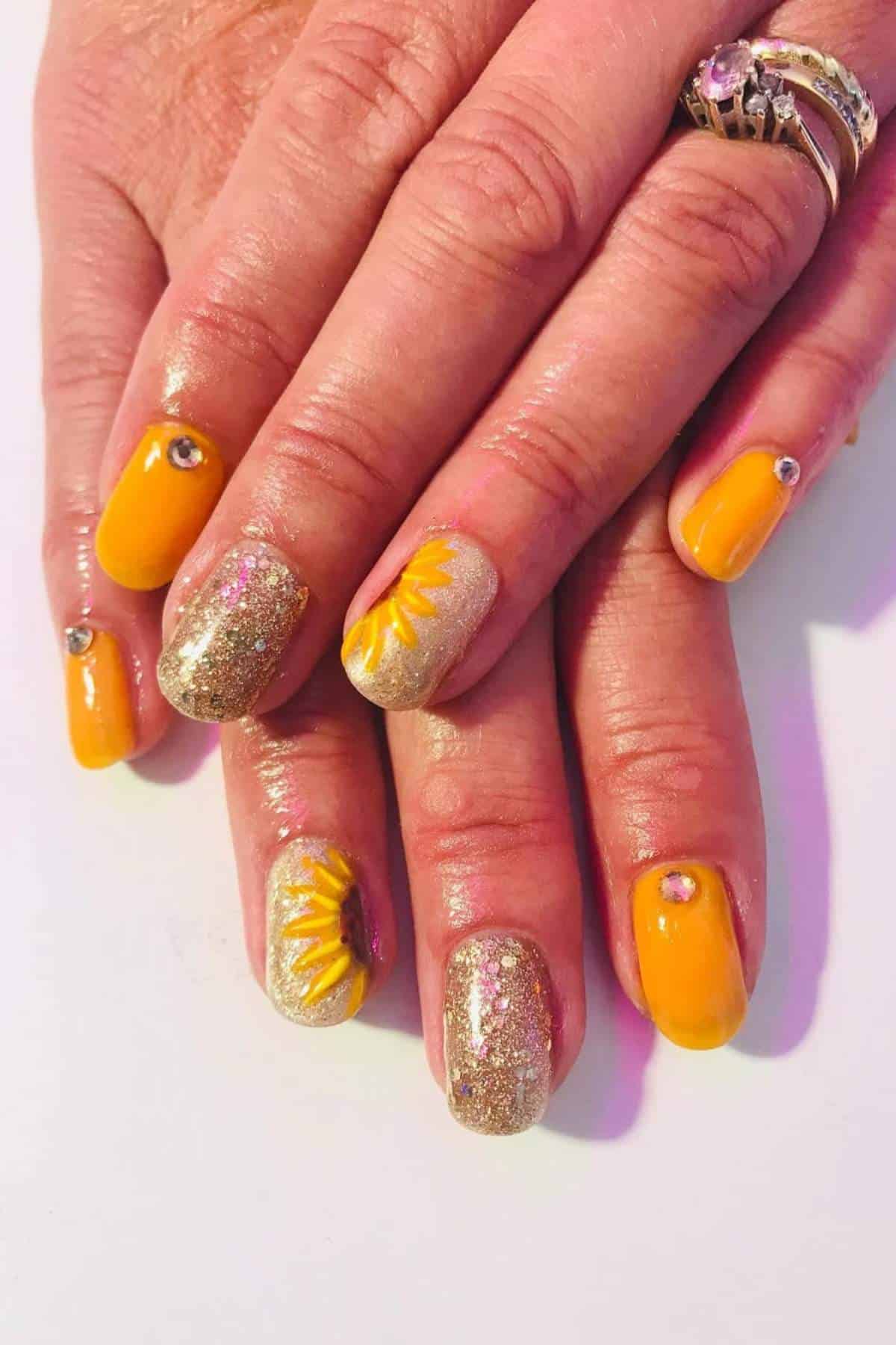 Hands with manicured nails painted yellow and gold glitter, accented with rhinestones and sunflower designs on two nails.