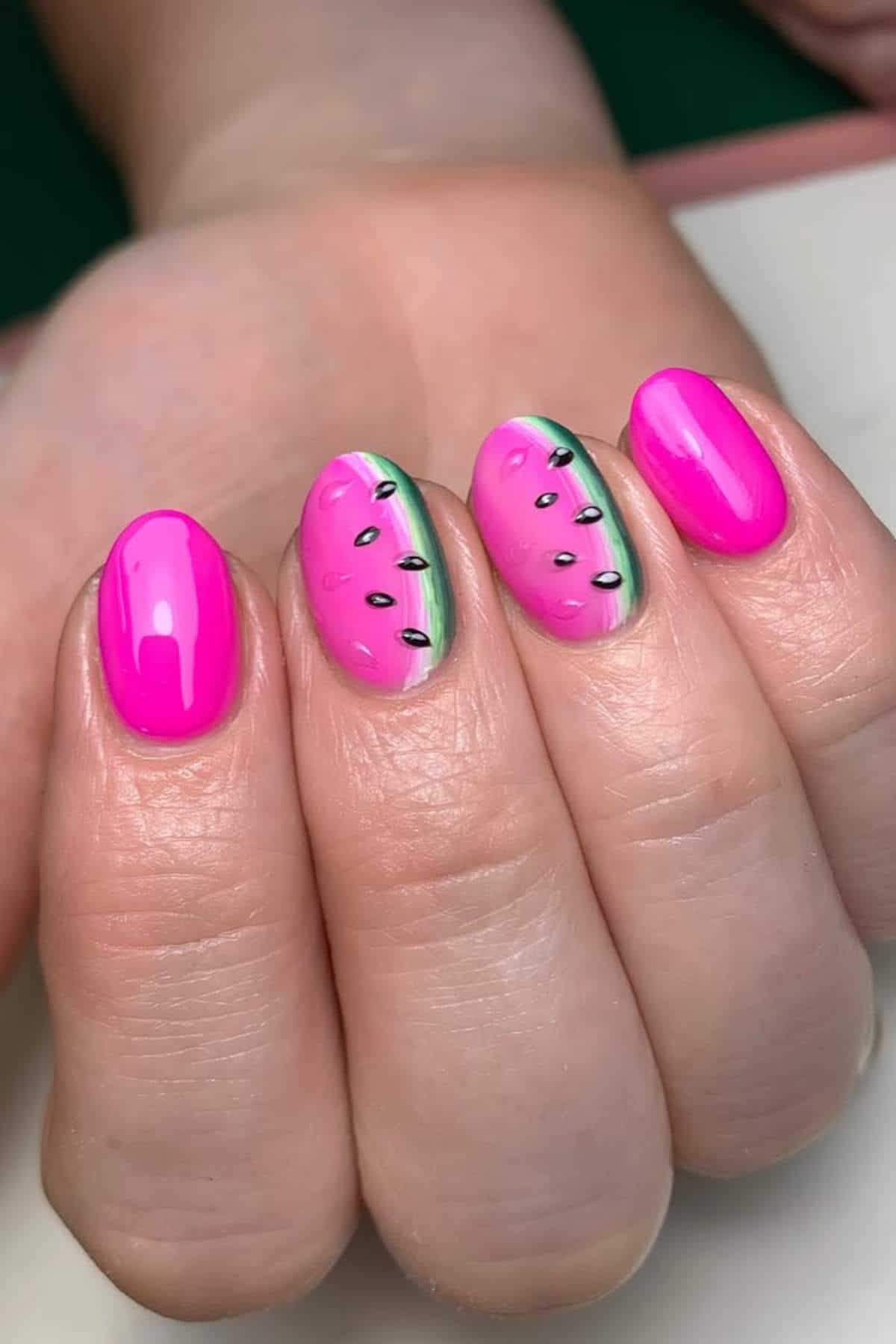 A hand with manicured nails; three nails are painted bright pink, and two nails are decorated with a watermelon design.