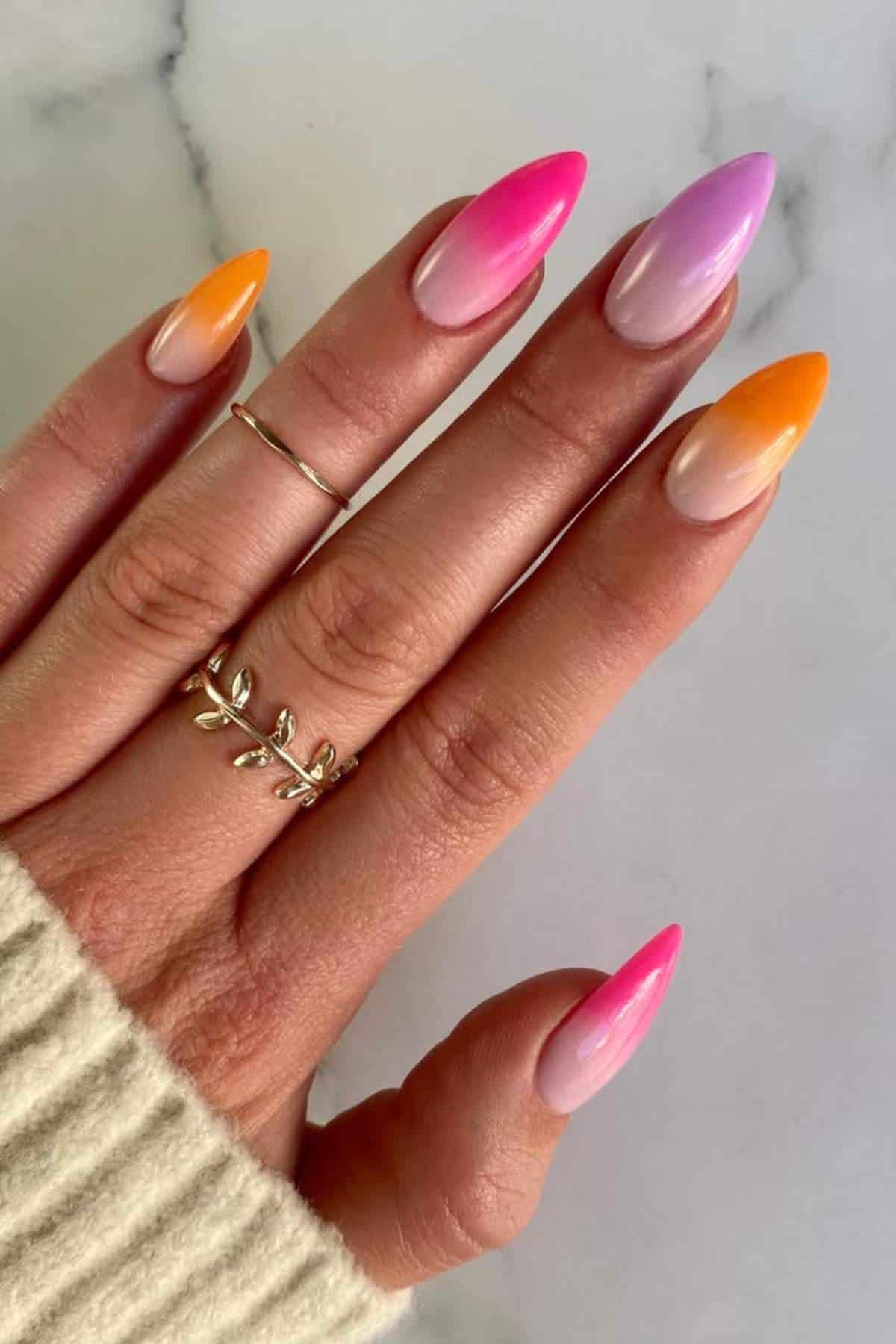 A hand with almond-shaped nails painted in a pink and orange ombre design, wearing two gold rings, shown against a white background.