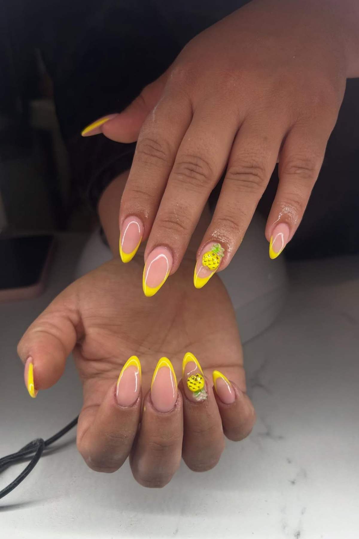 Two hands with long nails featuring yellow French tips; the ring finger on one hand has a yellow and black spotted accent design.