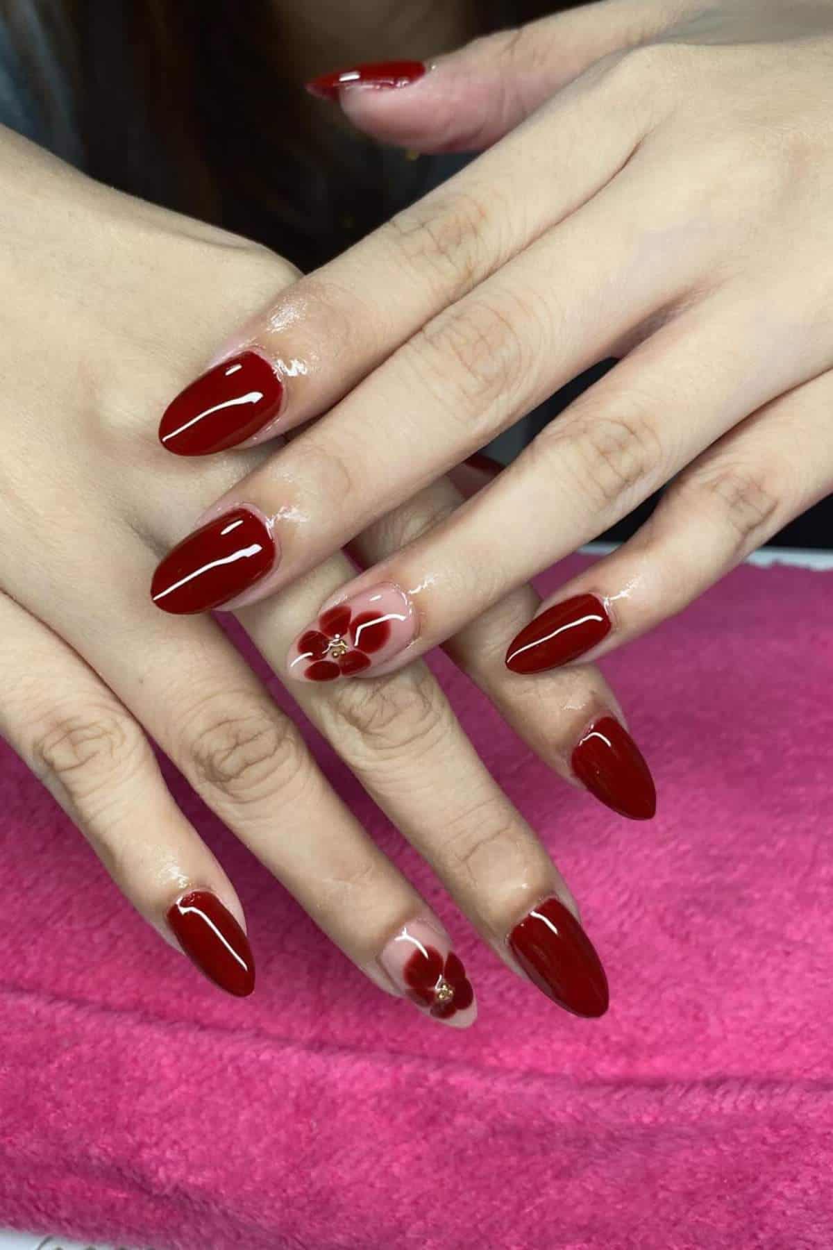 Hands with long, almond-shaped red nails; ring fingers feature a floral design on a nude base. Hands rest on a bright pink towel.