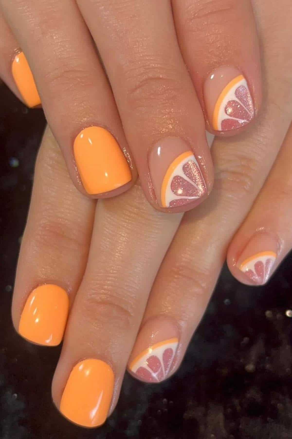 Hands with short, square-shaped nails painted orange, with two accent nails featuring a citrus fruit slice design in orange and glittery pink.