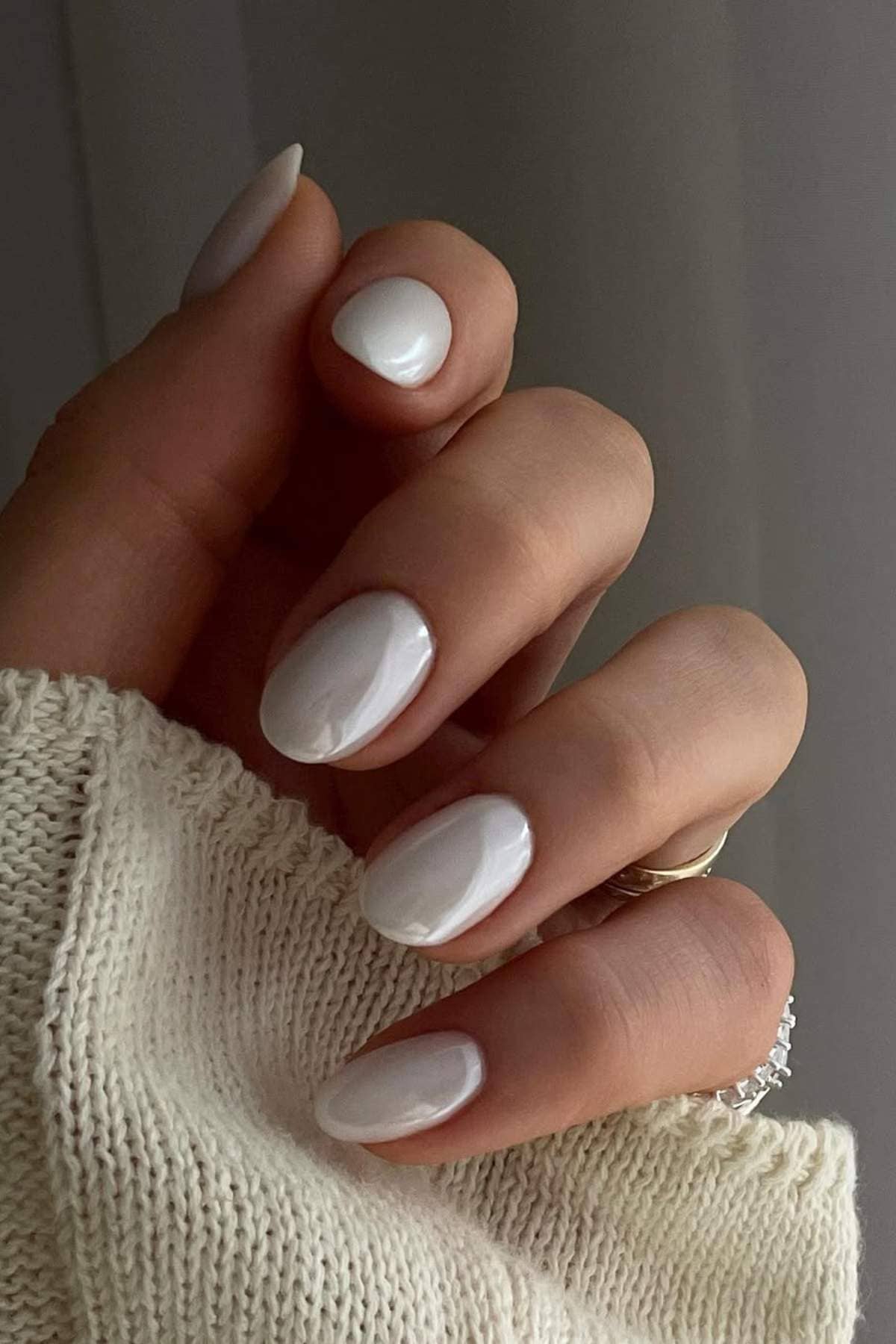A hand with short, almond-shaped nails painted glossy white, wearing a cream knit sleeve and two gold rings with small diamonds.