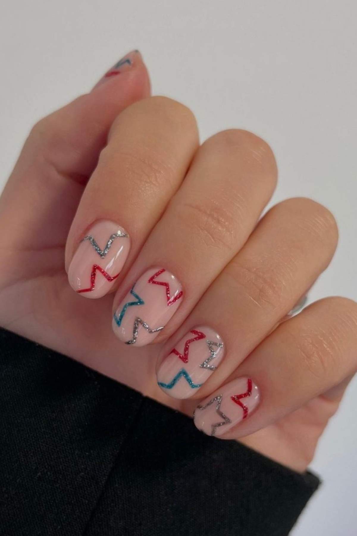 A hand with short nails painted in a nude polish, each nail decorated with wavy lines in red, silver, and blue.