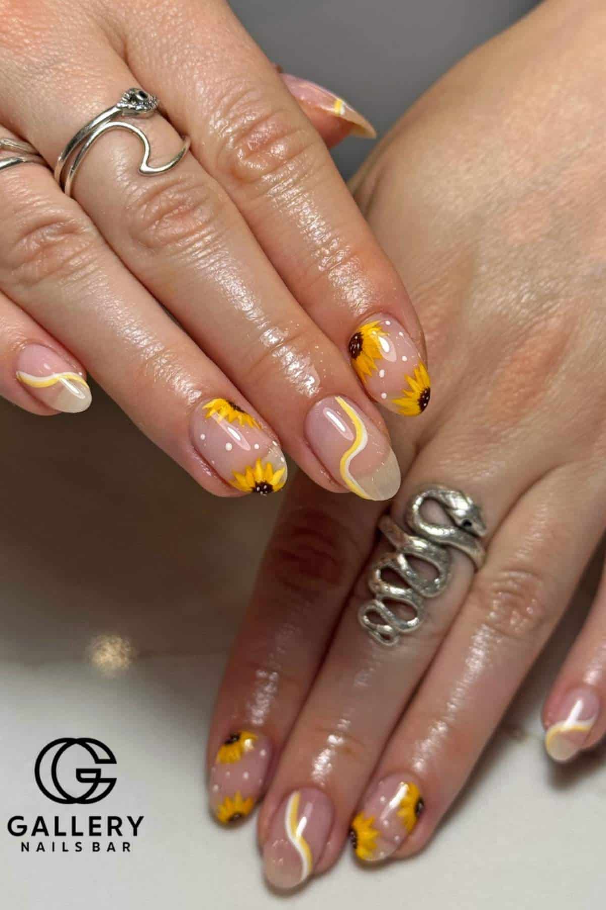Close-up of hands with manicured nails featuring sunflower designs, yellow and white wavy accents, and silver rings. "Gallery Nails Bar" logo is visible in the corner.