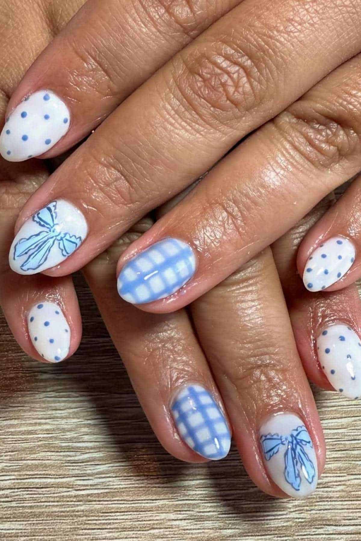 Fingernails with a light blue and white manicure featuring polka dots, plaid patterns, and delicate bow designs on a wooden surface.