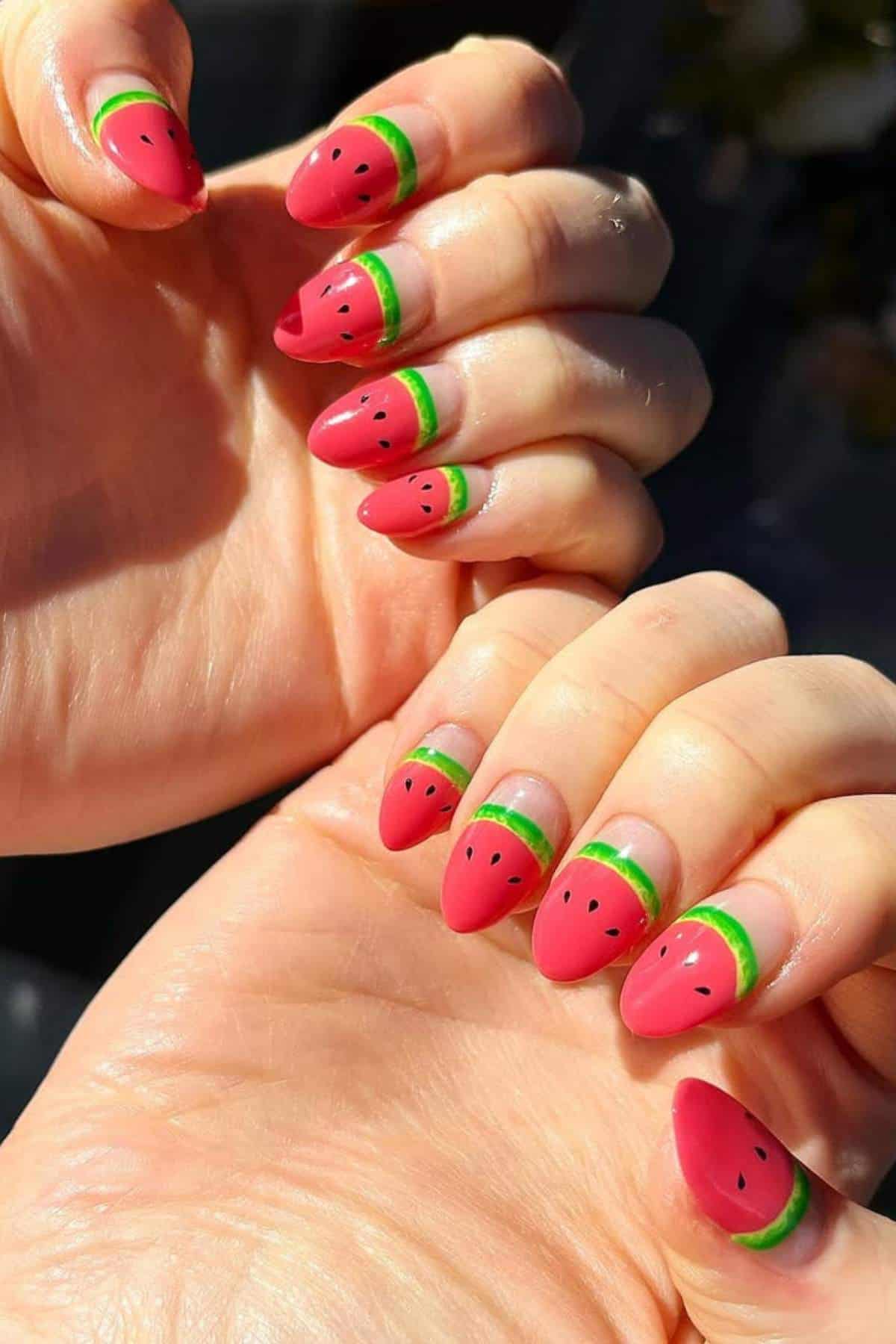 Hands with nails painted to resemble watermelon slices, featuring pink tips, green stripes, and small black seeds.