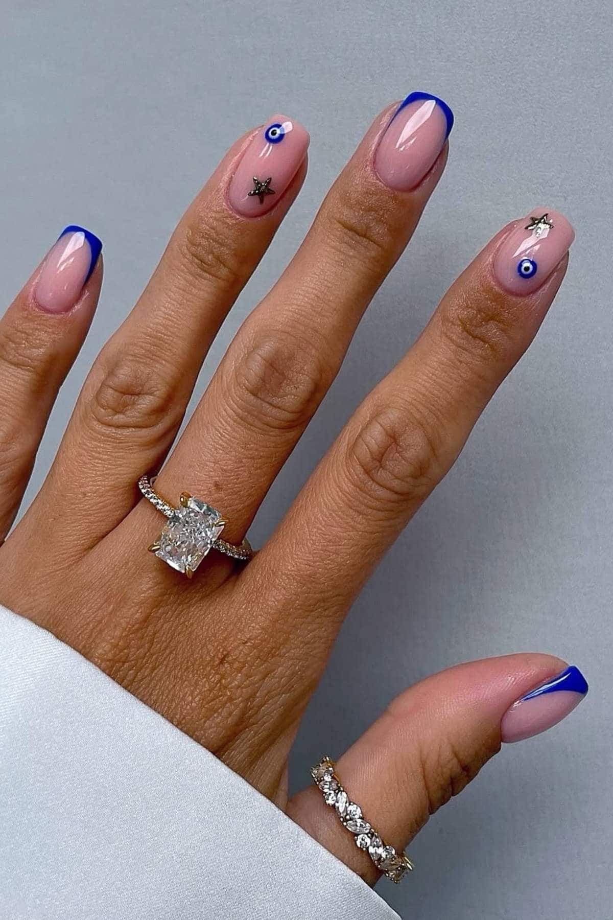A hand with pink nails featuring blue French tips and small nail art designs, wearing two diamond rings, against a light gray background.