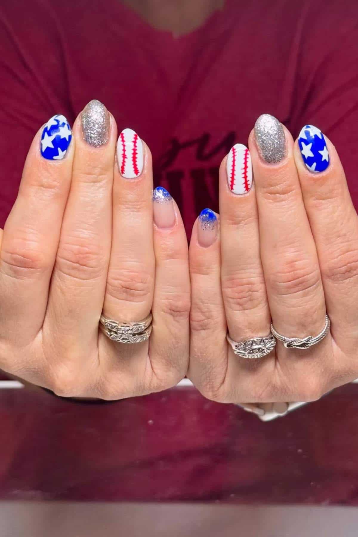 Hands with manicured nails featuring designs of blue stars, silver glitter, red baseball stitching, and blue French tips, with four silver rings on fingers.