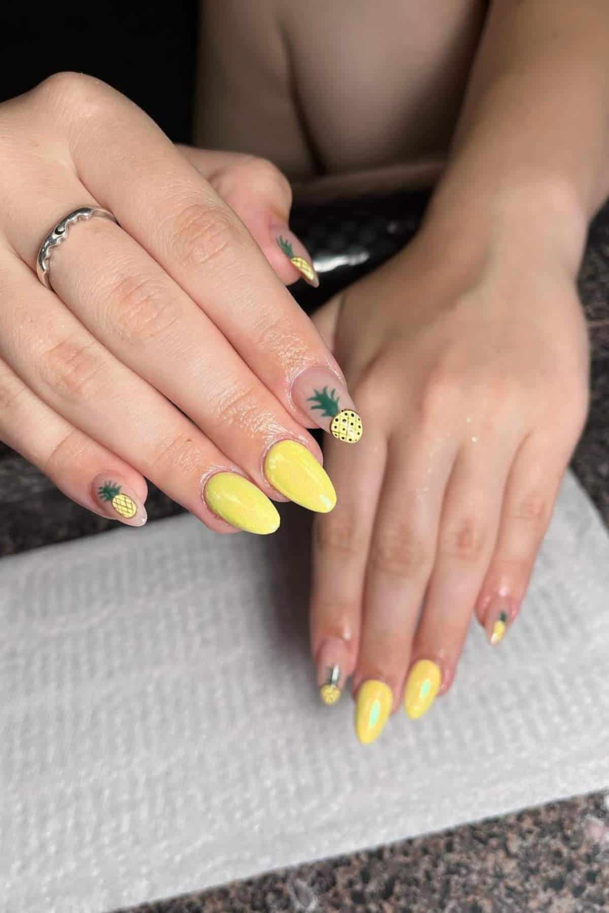 Hands with long, almond-shaped nails painted yellow and decorated with small pineapple designs, shown resting on a white paper towel on a granite surface.