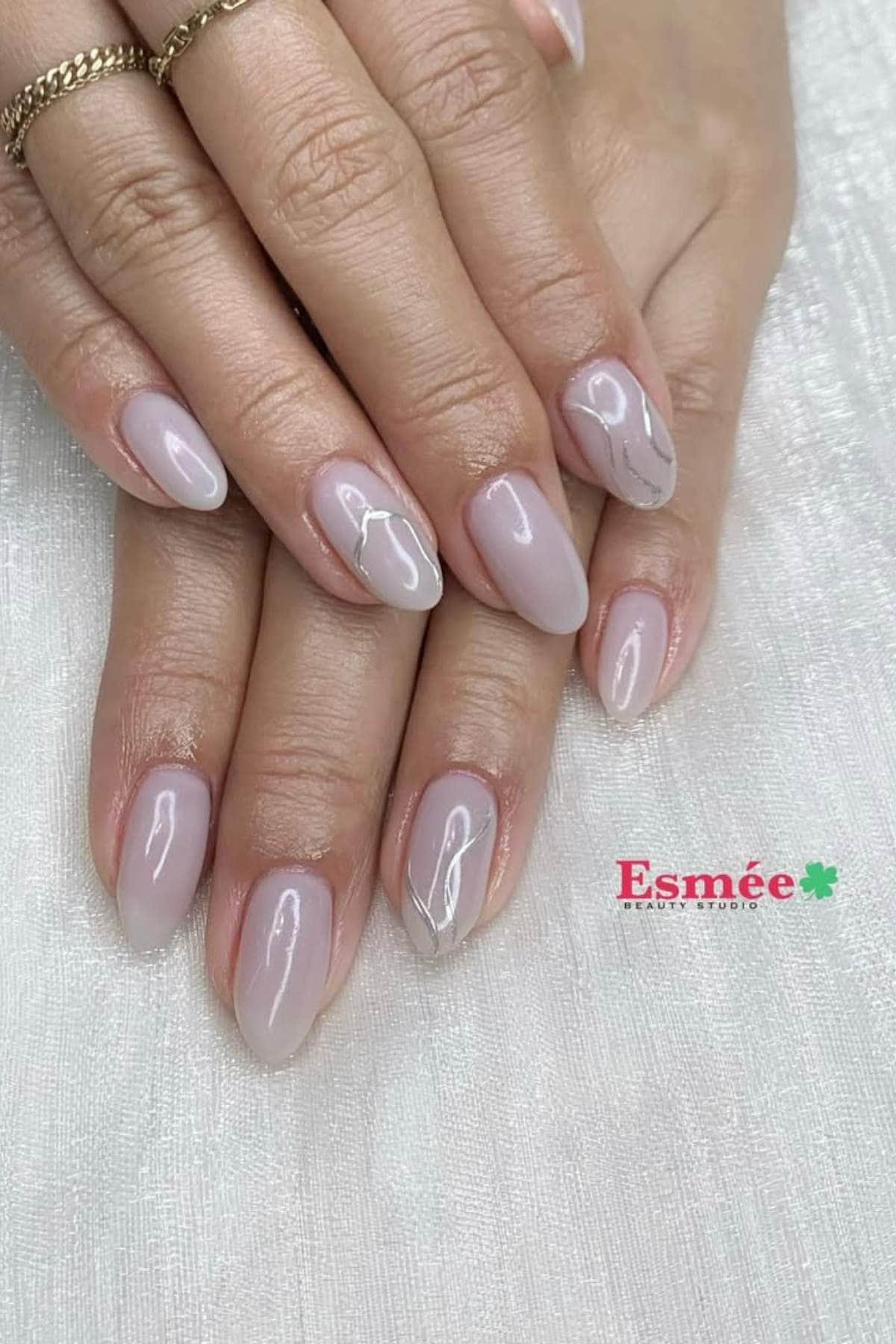 Hands with almond-shaped, pale pink nails featuring a glossy finish and subtle silver line accents on two fingers. The Esmée Beauty Studio logo is visible in the corner.